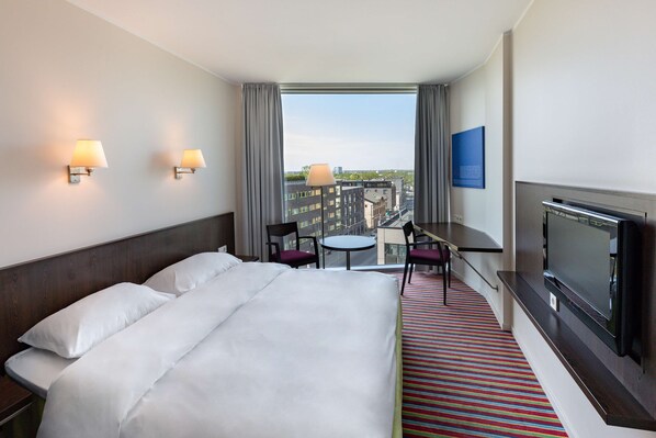 Una habitación de hotel con una cama, dos lámparas de pared, un televisor, una mesita con sillas y vistas a la ciudad a través de amplios ventanales.