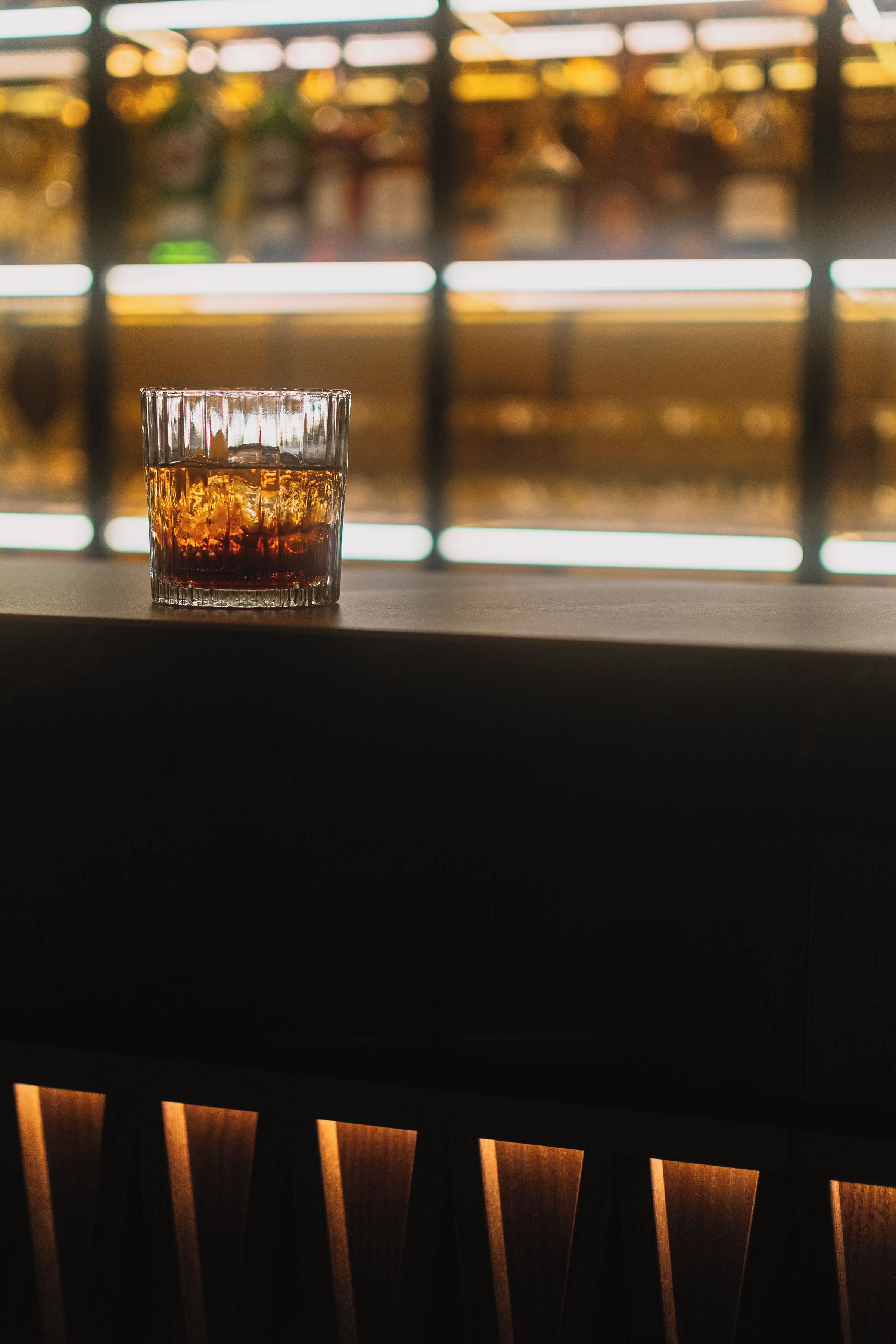 A glass of whiskey on a bar counter with a blurred background of shelves and bottles.