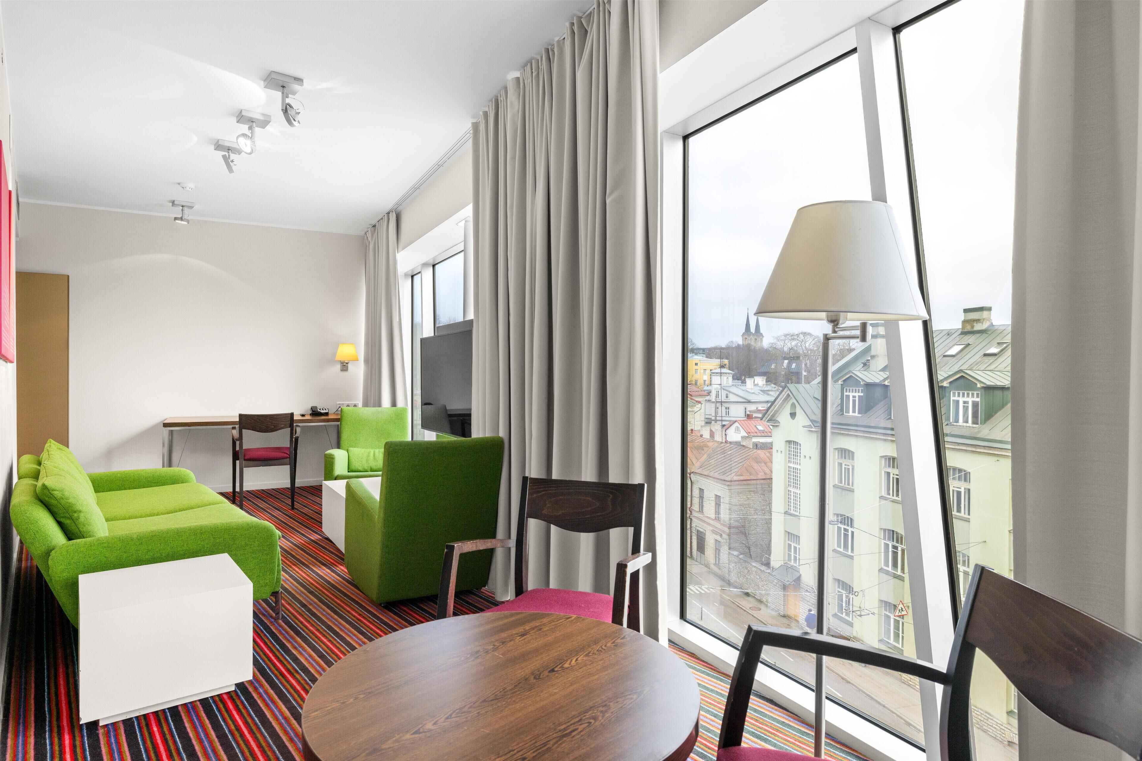 Habitación de hotel con un ventanal grande, una mesa de comedor de madera, sofás blancos y verdes, y un televisor.