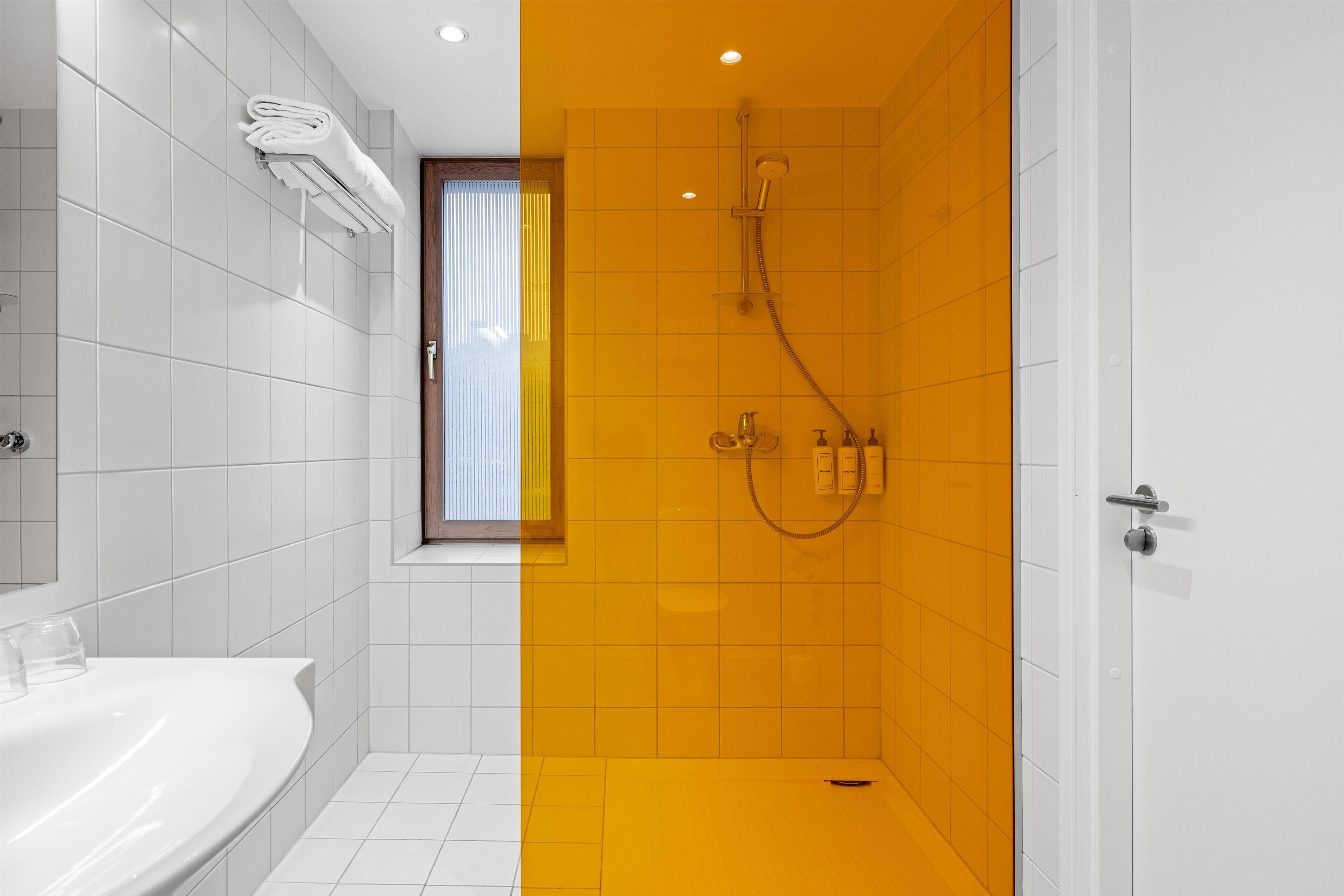 A modern bathroom with white and yellow tiles, a built-in shower, and a window with blinds.