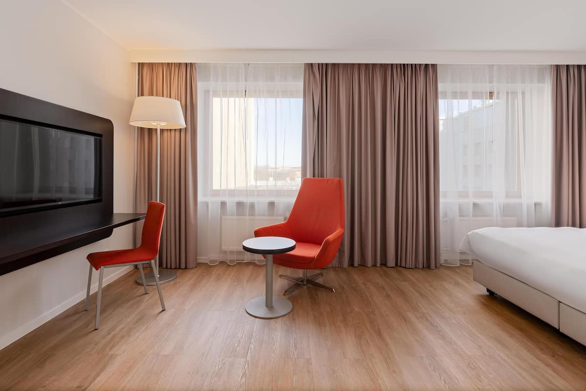 Ein Hotelzimmer mit Bett, Fernseher, Schreibtisch mit Stuhl, kleinem Tisch und einem Fenster mit Vorhängen.