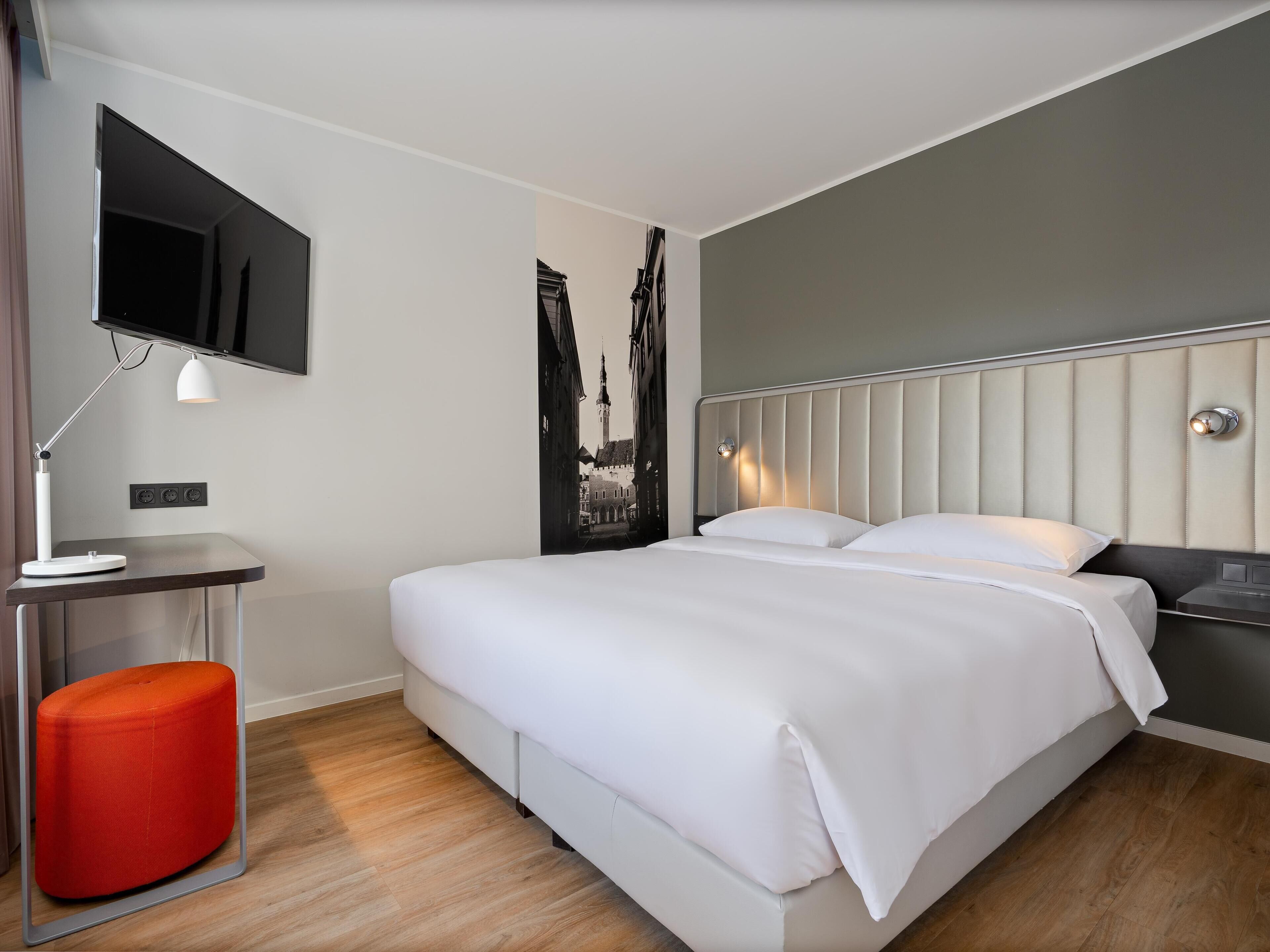 A hotel room with a bed, a desk, a chair, a TV, and a wall-mounted light.