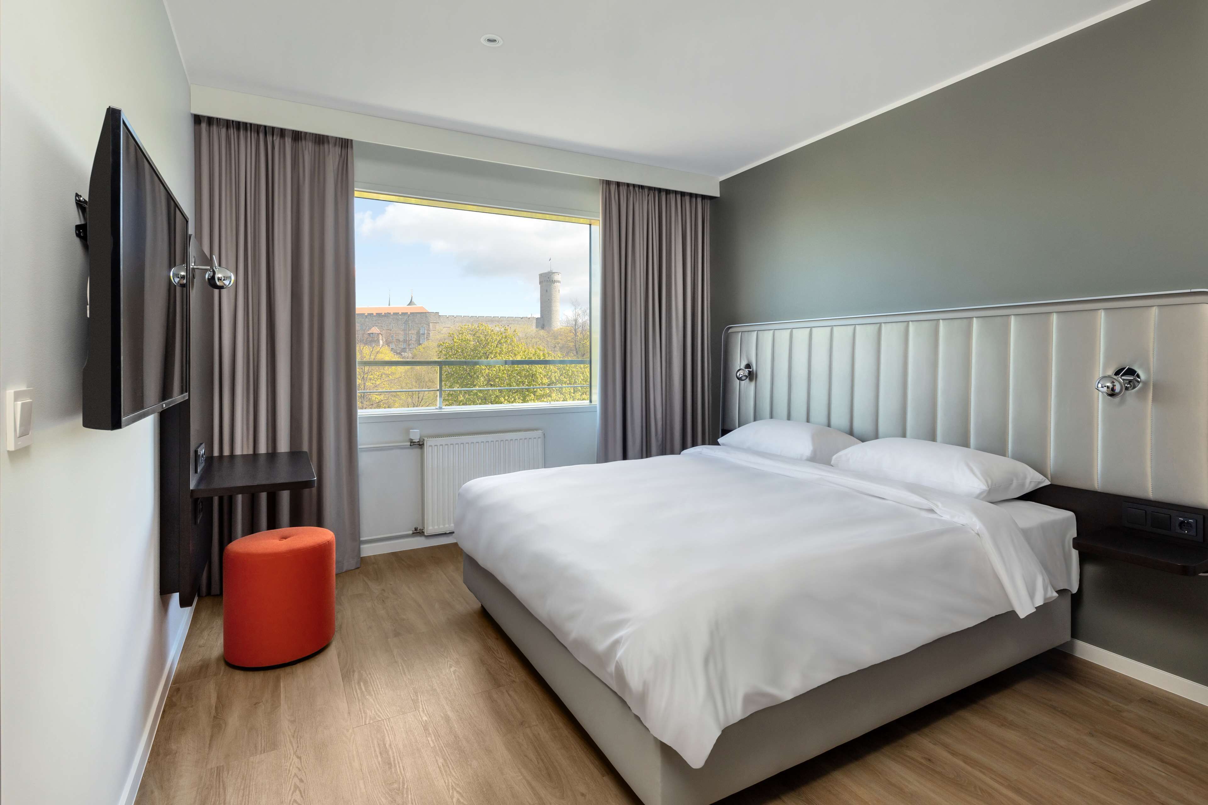 A modern hotel room with a large bed, a TV mounted on the wall, a window with curtains, and a red ottoman.