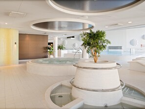 Un área de piscina interior moderna con un jacuzzi central, rodeado de asientos y una escalera que conduce a un nivel inferior.