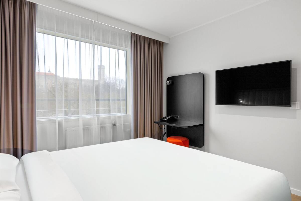 Ein Hotelzimmer mit einem großen Bett, einem an der Wand montierten Fernseher, einem kleinen Tisch und einem Fenster mit Vorhängen.