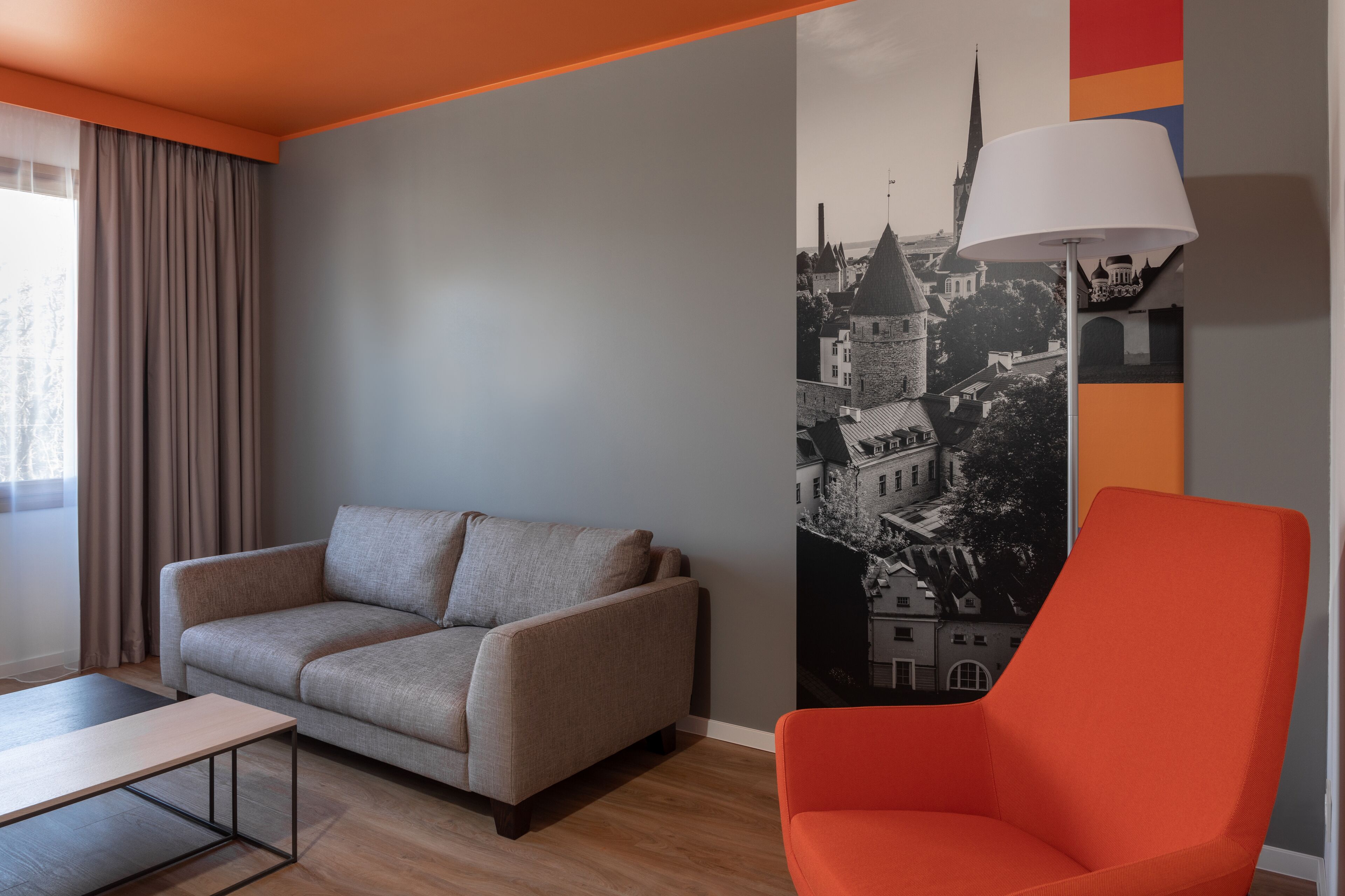 Una sala moderna con un sofá gris, un sillón rojo, una mesa blanca y un mural de un paisaje urbano en la pared.