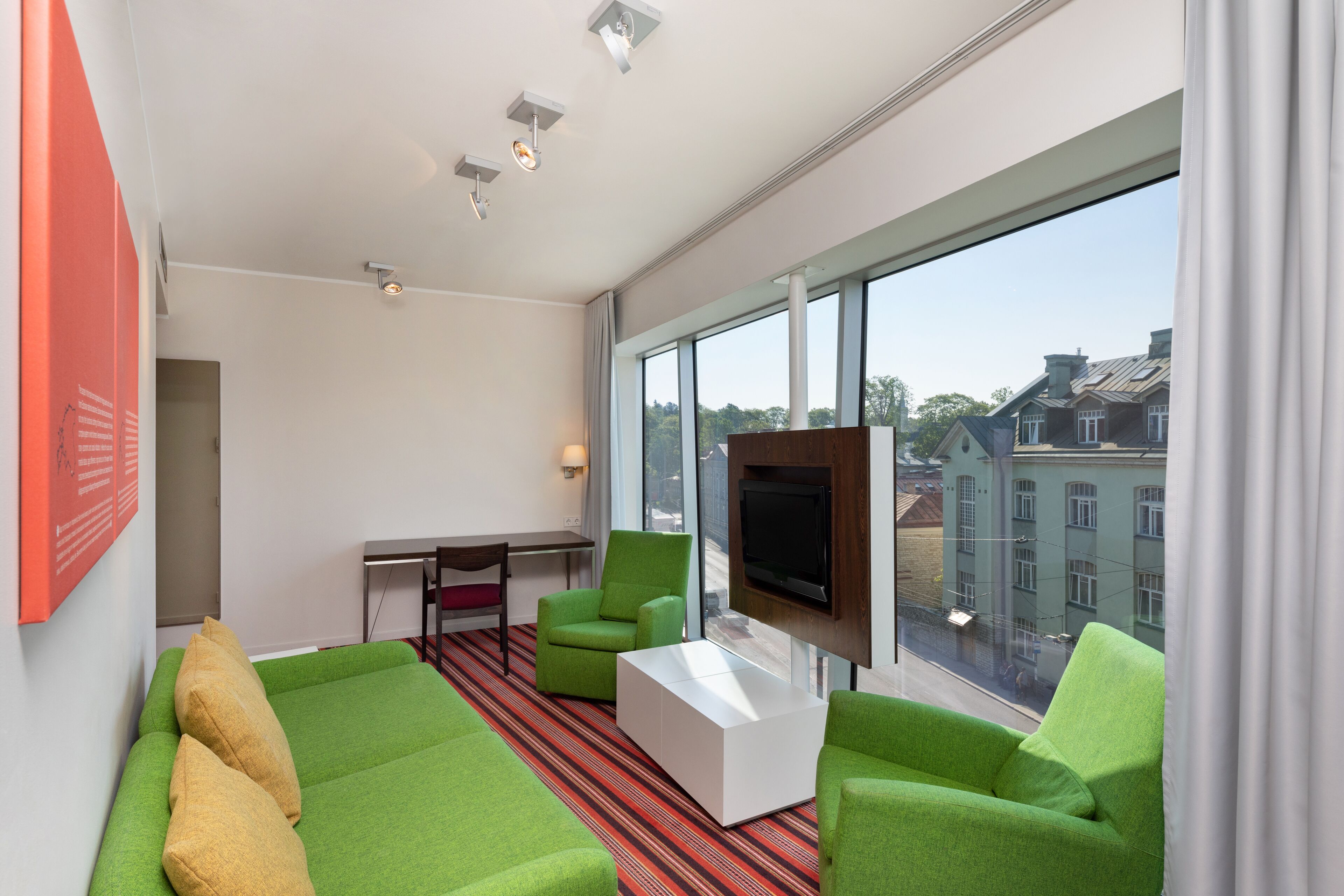 A modern hotel room with a green sofa, a white coffee table, and a flat-screen TV mounted on the wall.