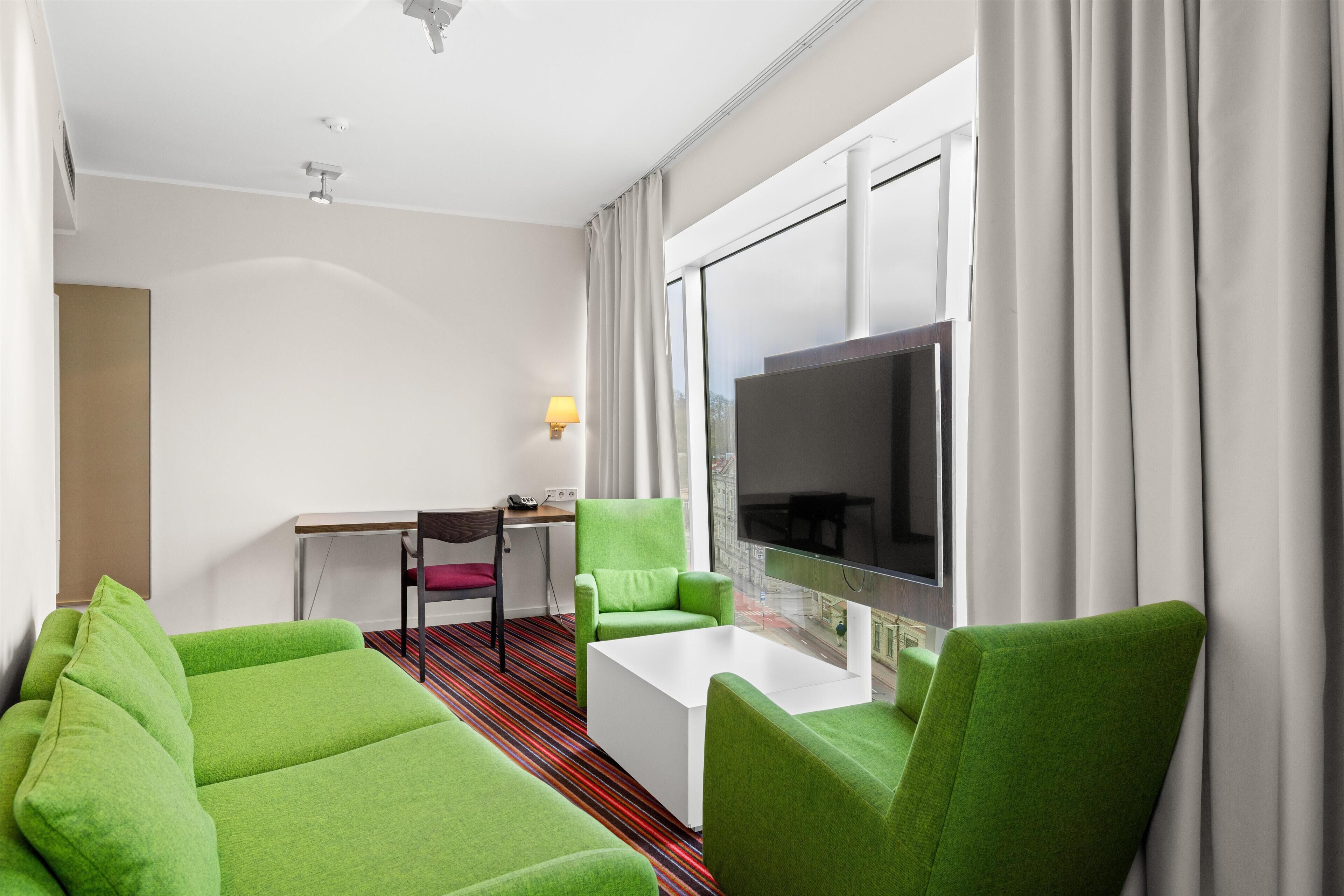 Una habitación de hotel moderna con un sofá verde, una mesa de centro blanca, un televisor de pantalla plana y un escritorio con lámpara.