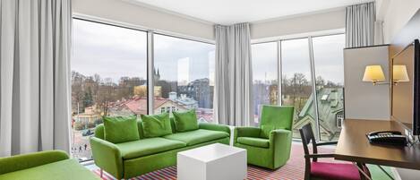 Una habitación de hotel moderna con ventanas amplias, un sofá verde, una mesa de centro blanca y una alfombra a rayas.