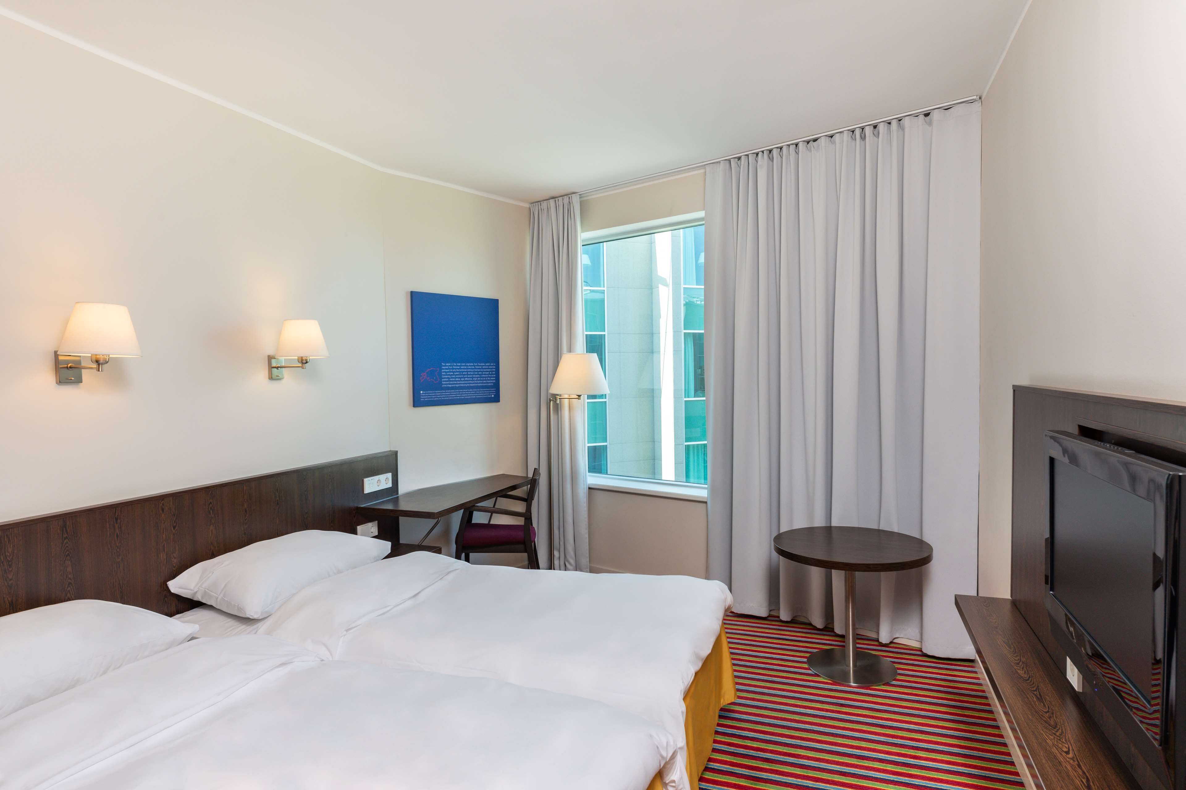 Une chambre d’hôtel avec un grand lit, un bureau, une chaise, une petite table, un téléviseur et une fenêtre avec des rideaux.