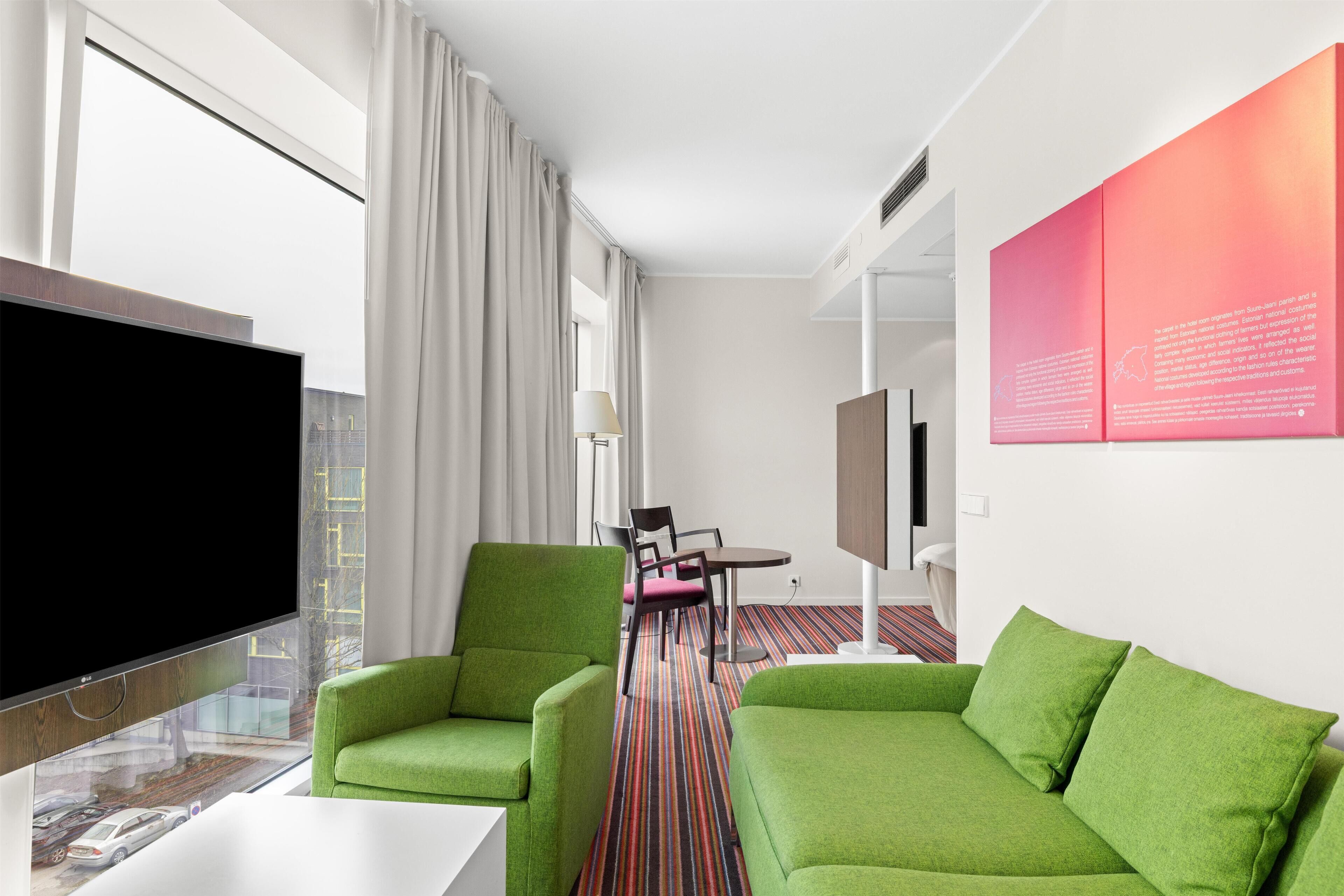 A hotel room with a large flat-screen TV, green upholstered armchairs, a small round table, and a colorful striped carpet.