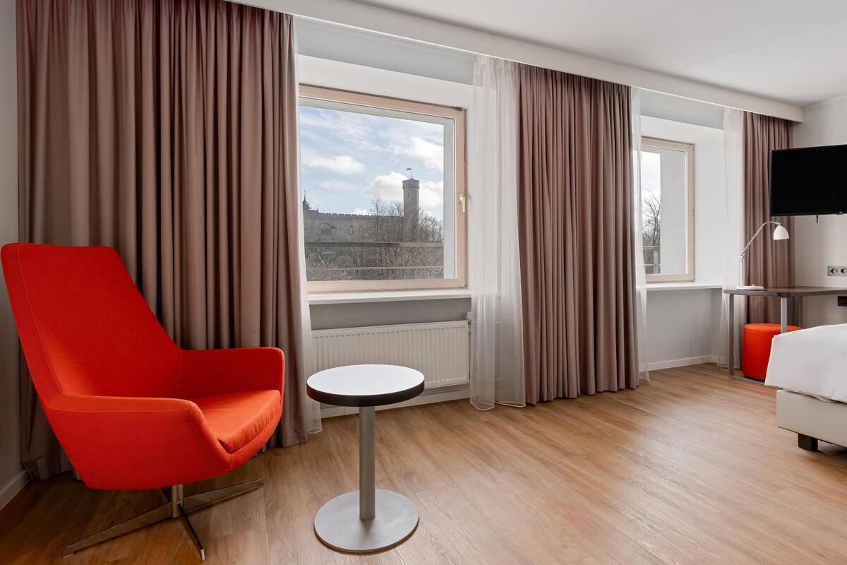 Ein Hotelzimmer mit rotem Sessel, kleinem runden Tisch, Bett, Fernseher und einem Fenster mit Vorhängen.