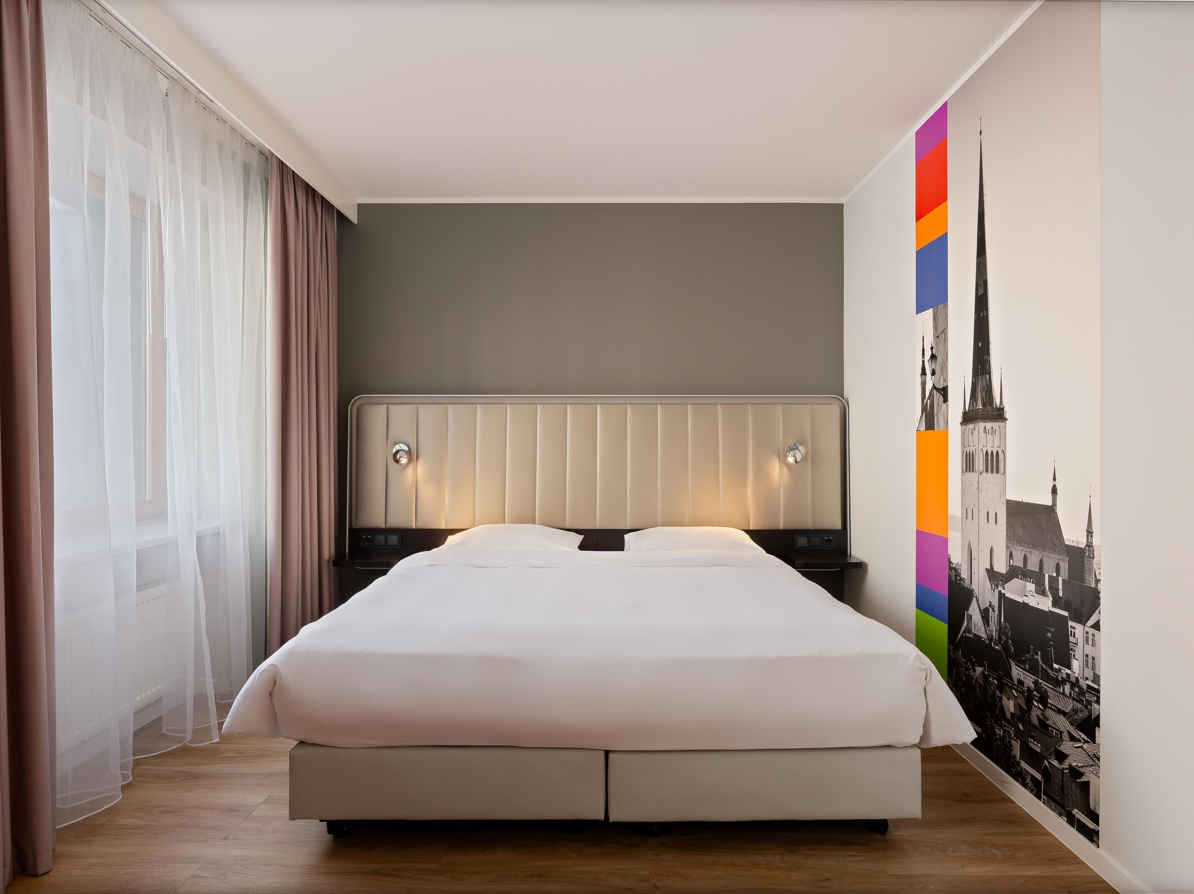 A modern hotel room with a large bed, two bedside tables, and a cityscape wall art.