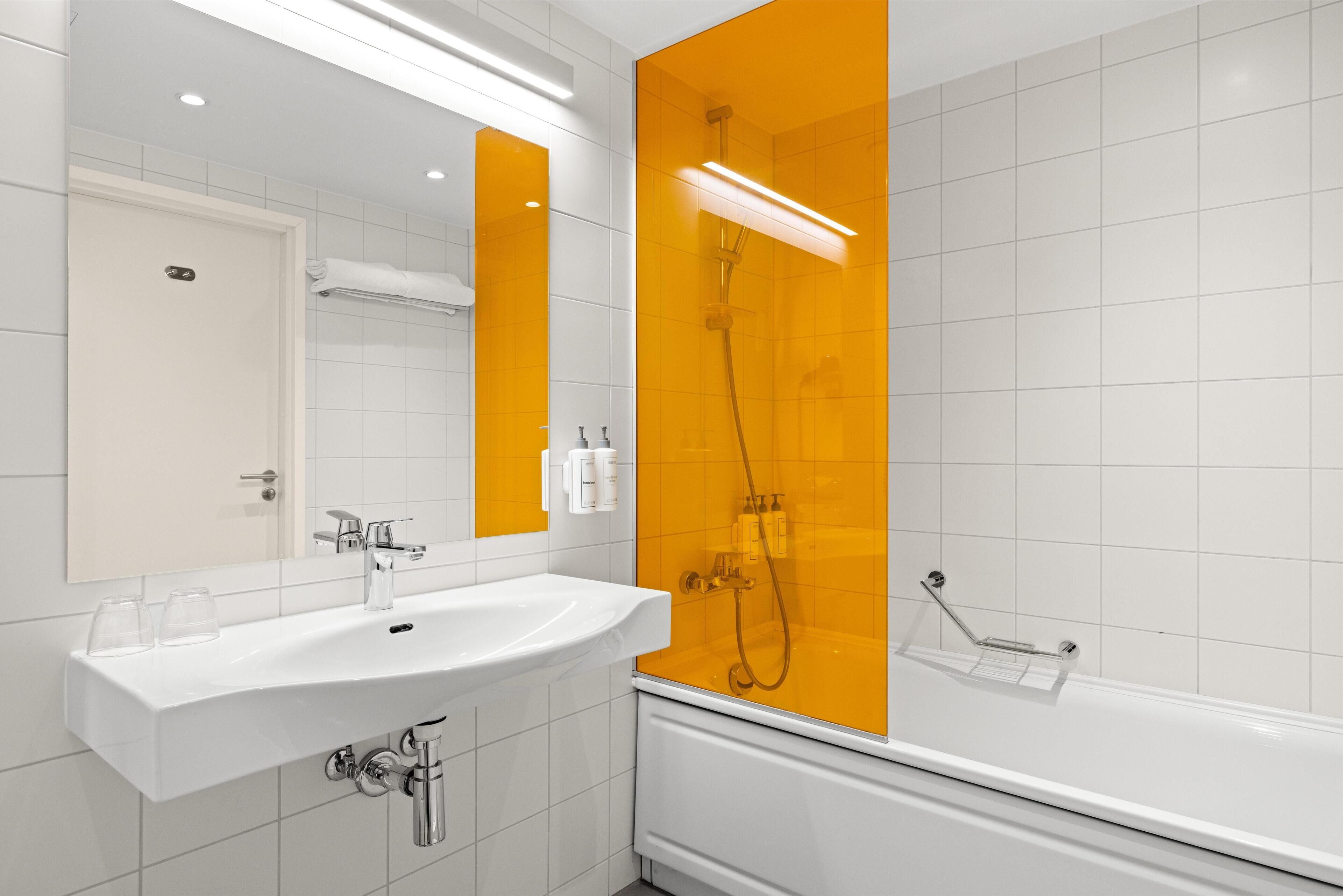 Un baño moderno con un lavamanos blanco, espejo y ducha con una pared de acento amarillo.