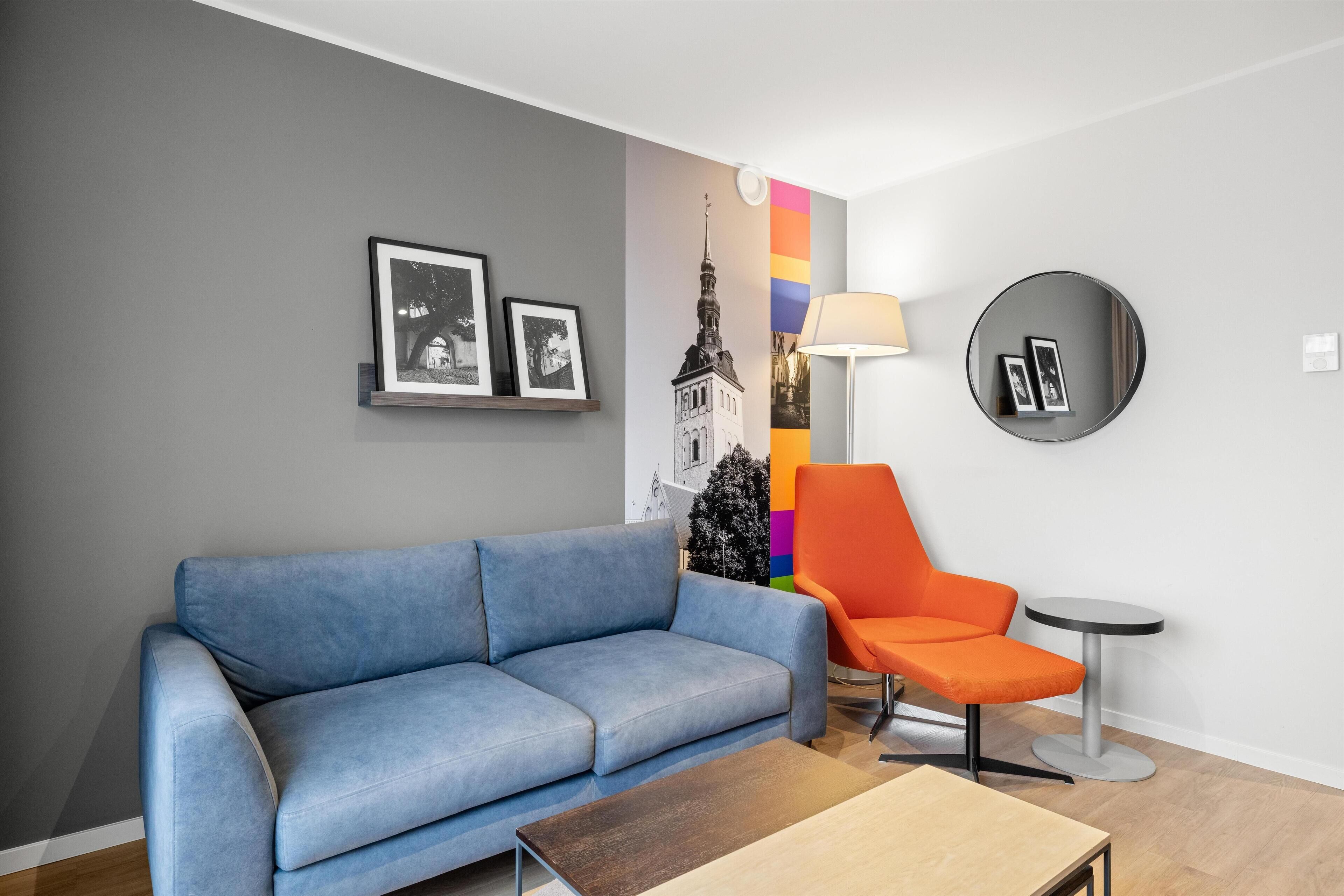Una sala de estar moderna con un sofá azul, un sillón naranja, un espejo redondo y una pequeña mesa circular.