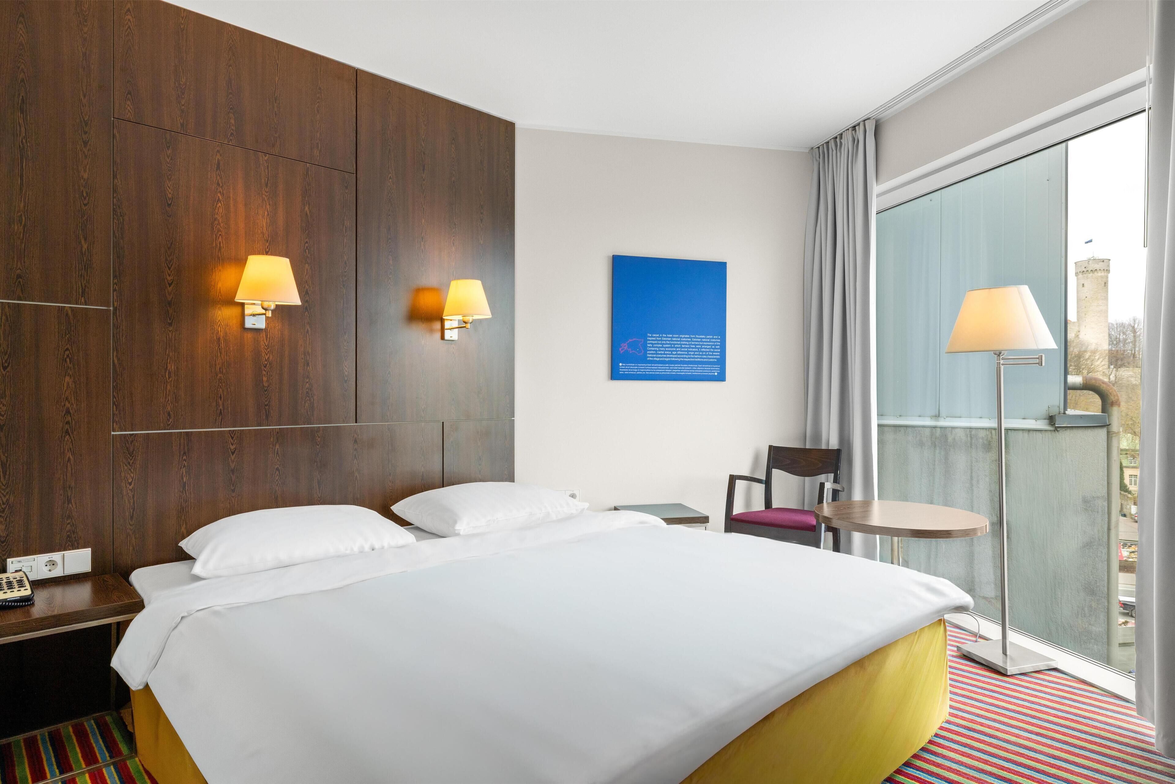 A hotel room with a large bed, two wall-mounted lamps, a small table, a chair, and a view of a tower through a large window.