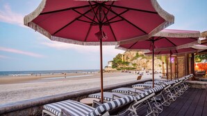 Une terrasse au bord de la plage, dotée de chaises longues à rayures et de parasols rouges, qui offre une vue sur une plage de sable et des falaises rocheuses.