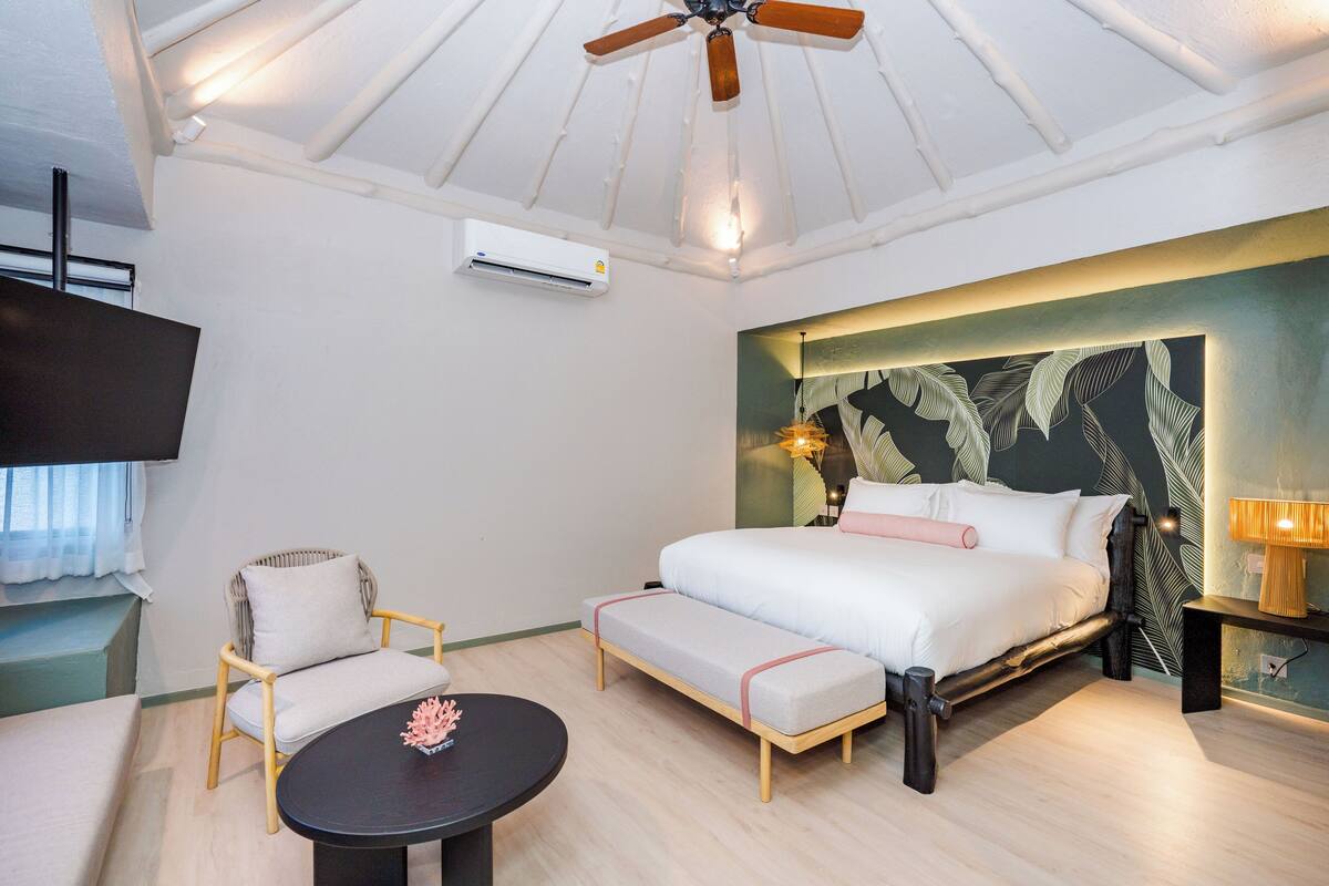 Une chambre à coucher comprenant un lit, une chaise, une petite table, un ventilateur de plafond et un téléviseur fixé au mur.