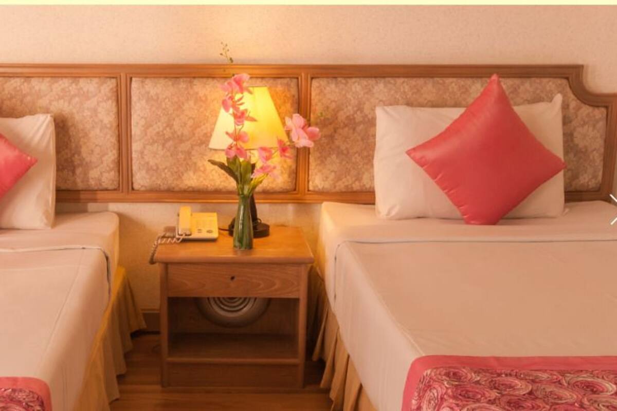Une chambre d’hôtel avec deux lits, chacun doté d’une tête de lit en bois et d’oreillers roses. Une table de chevet se trouve entre les deux lits, sur laquelle reposent une lampe, un vase avec des fleurs et un téléphone.