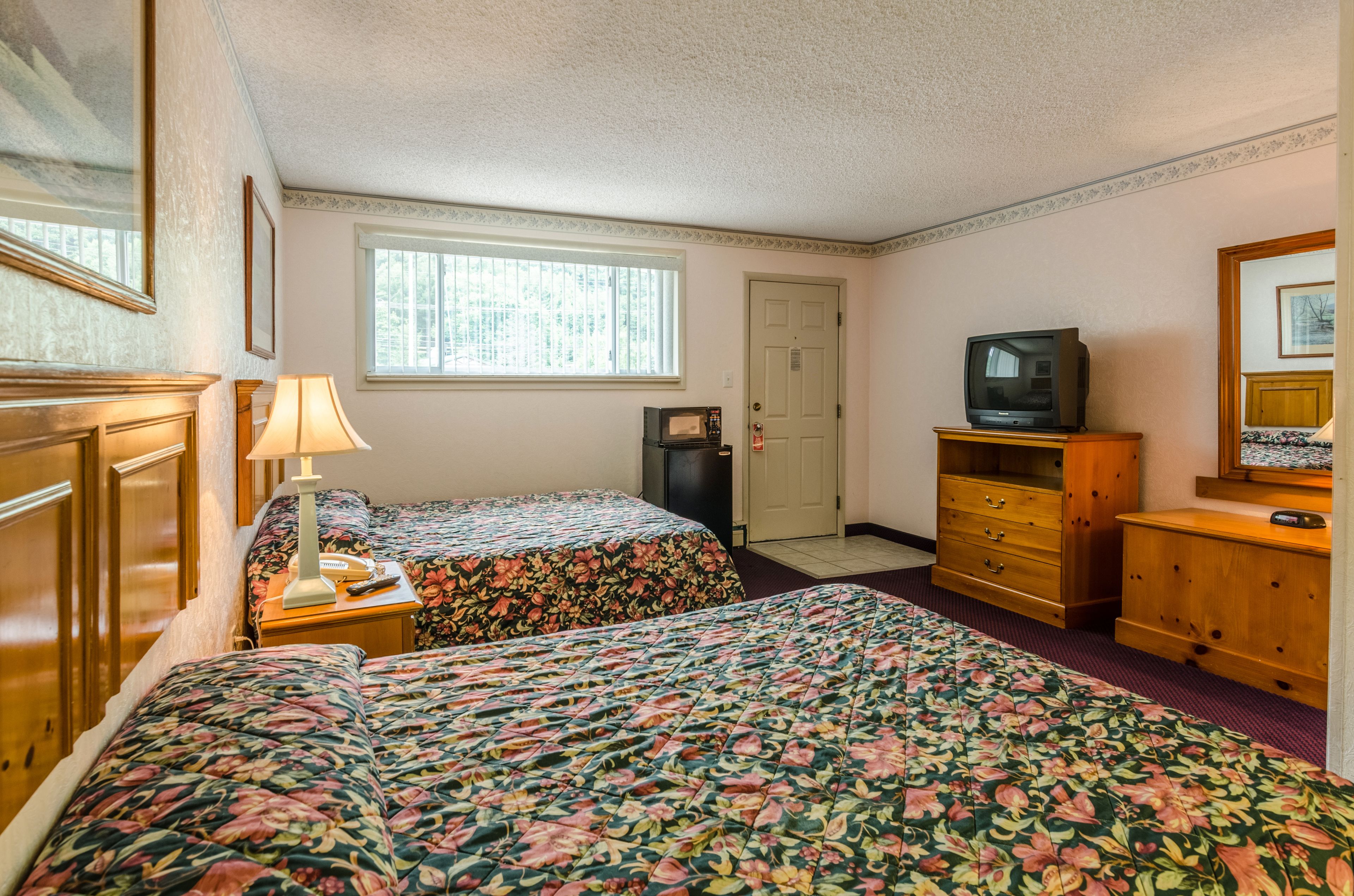 A hotel room with two beds, a television, a dresser, and a window with blinds.