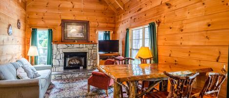 A log cabin interior with a stone fireplace, wooden dining table, and a television.