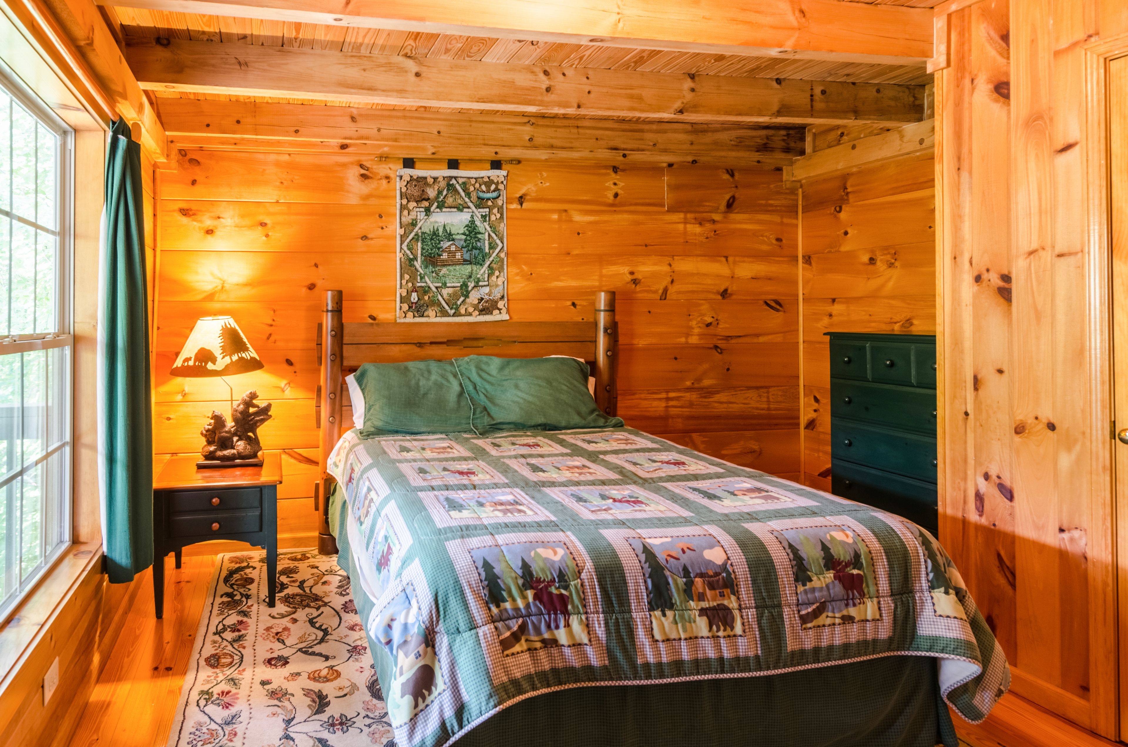 Un dormitorio rústico de cabaña de madera, con una cama, una mesita de noche con lámpara, un aparador verde y una ventana con cortinas.