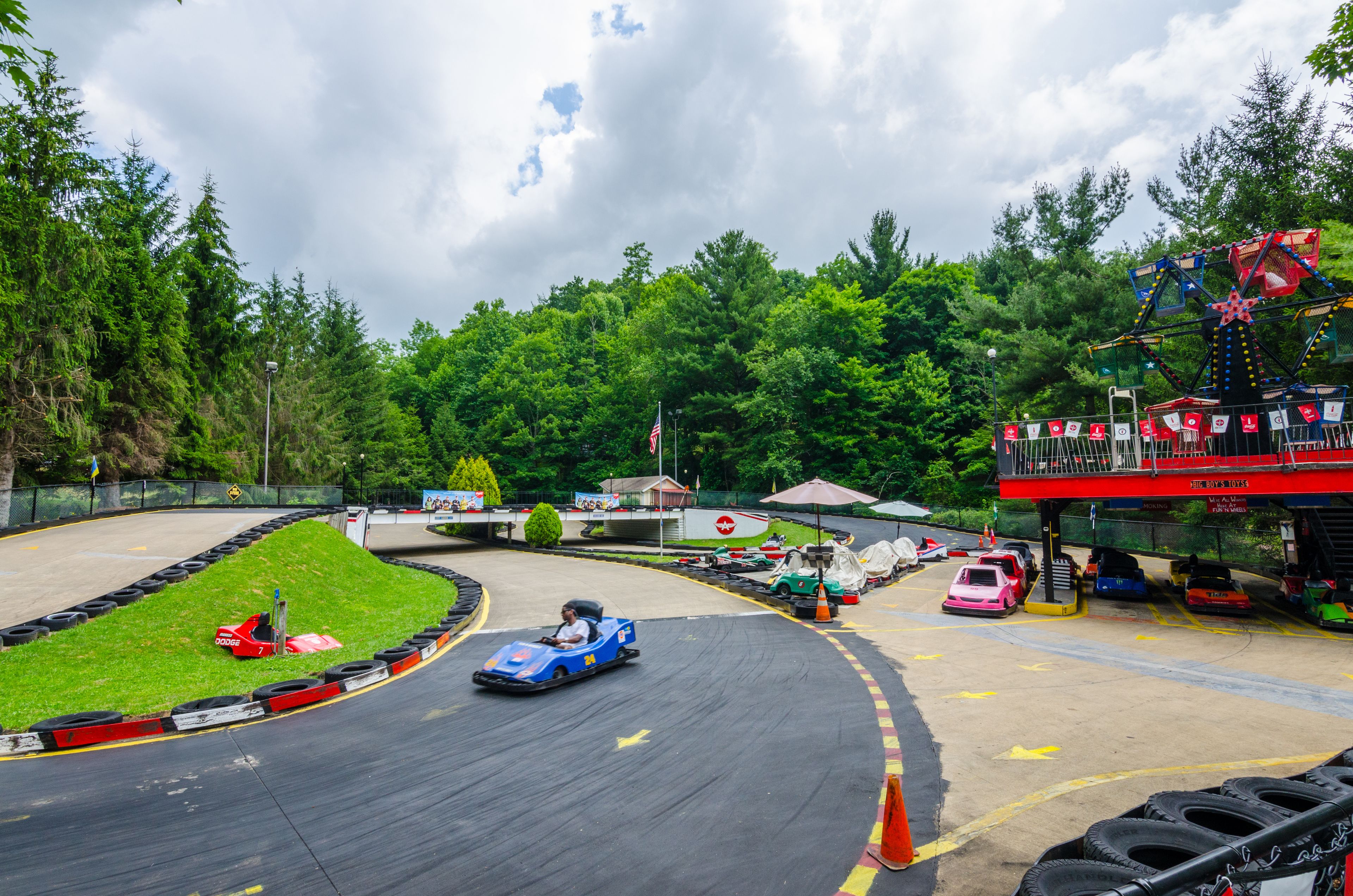 A go-kart track with a blue go-kart in motion, surrounded by greenery and a Ferris wheel in the background.