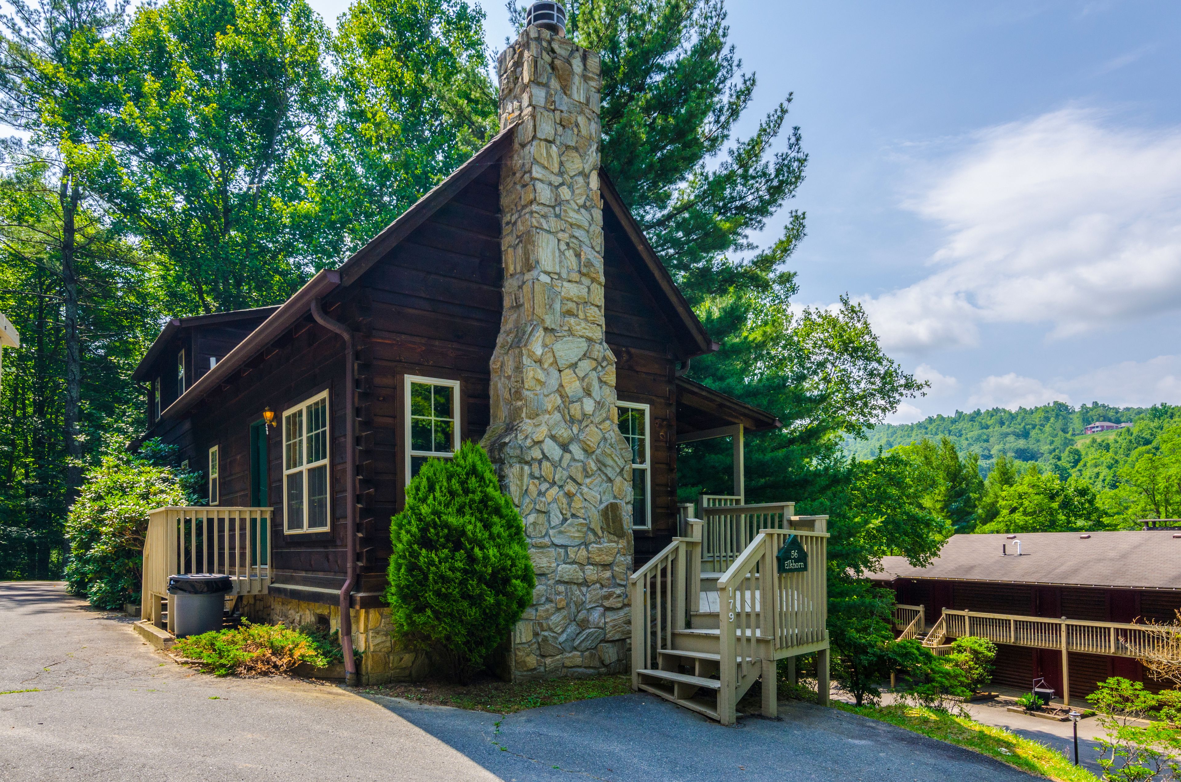 A rustic cabin with a stone chimney, wooden deck, and a view of the forest.