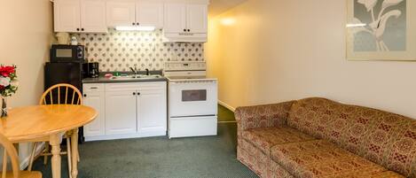 A compact kitchen with white cabinets, a microwave, a stove, and a sink. There is a patterned sofa and a dining table with chairs.