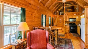 Ein gemütliches Blockhaus-Interieur mit Essbereich, einem rot gepolsterten Sessel, einem Holztisch, einer Küche mit Geräte und einem großen Fenster mit Vorhängen.