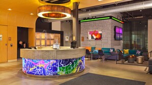 Un área de recepción moderna con un mostrador de recepción iluminado y colorido, un espacio de descanso con sofás y sillones, y una gran pantalla en la pared.