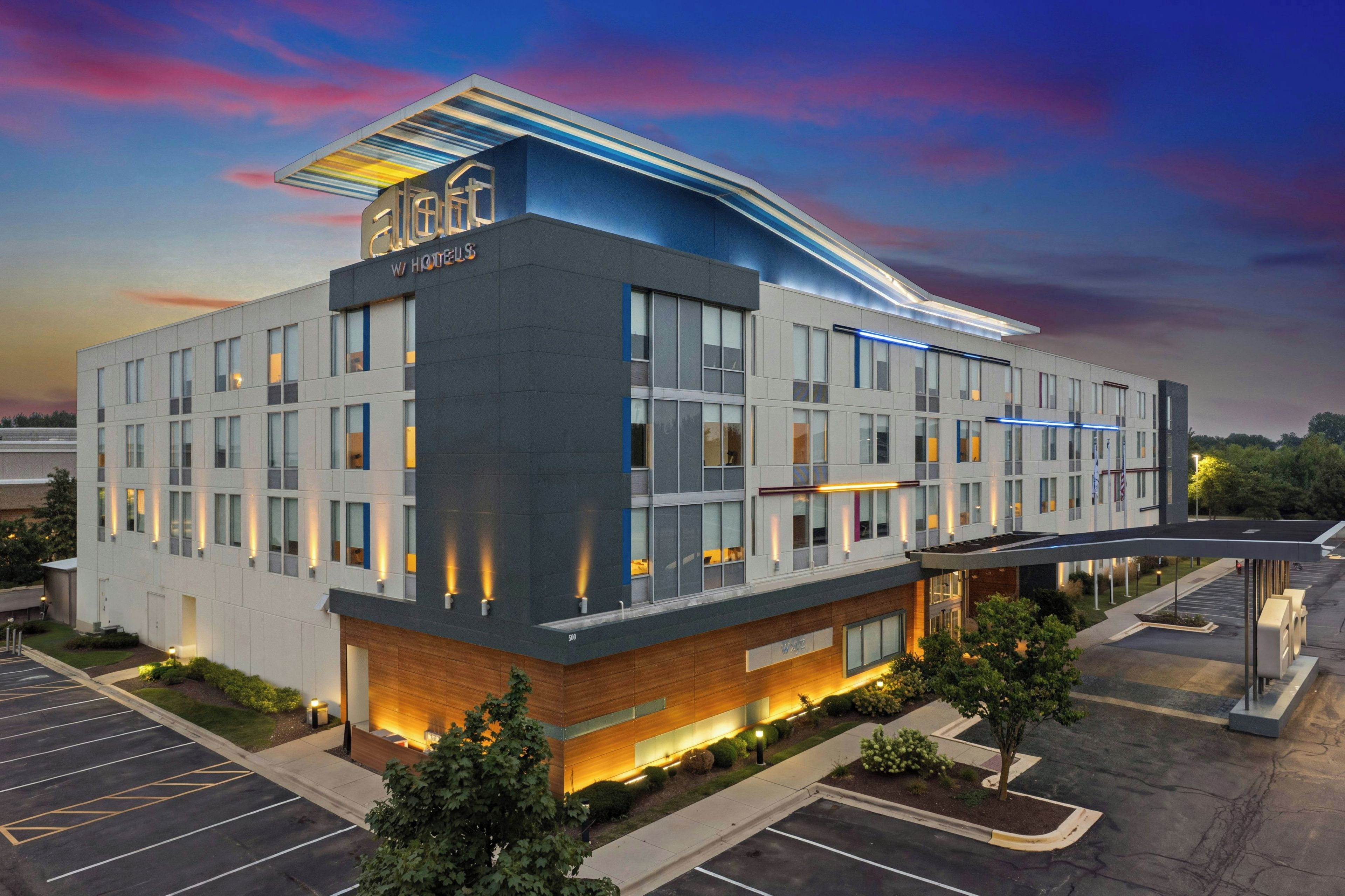 A modern hotel building with a flat roof, large windows, and a parking lot.