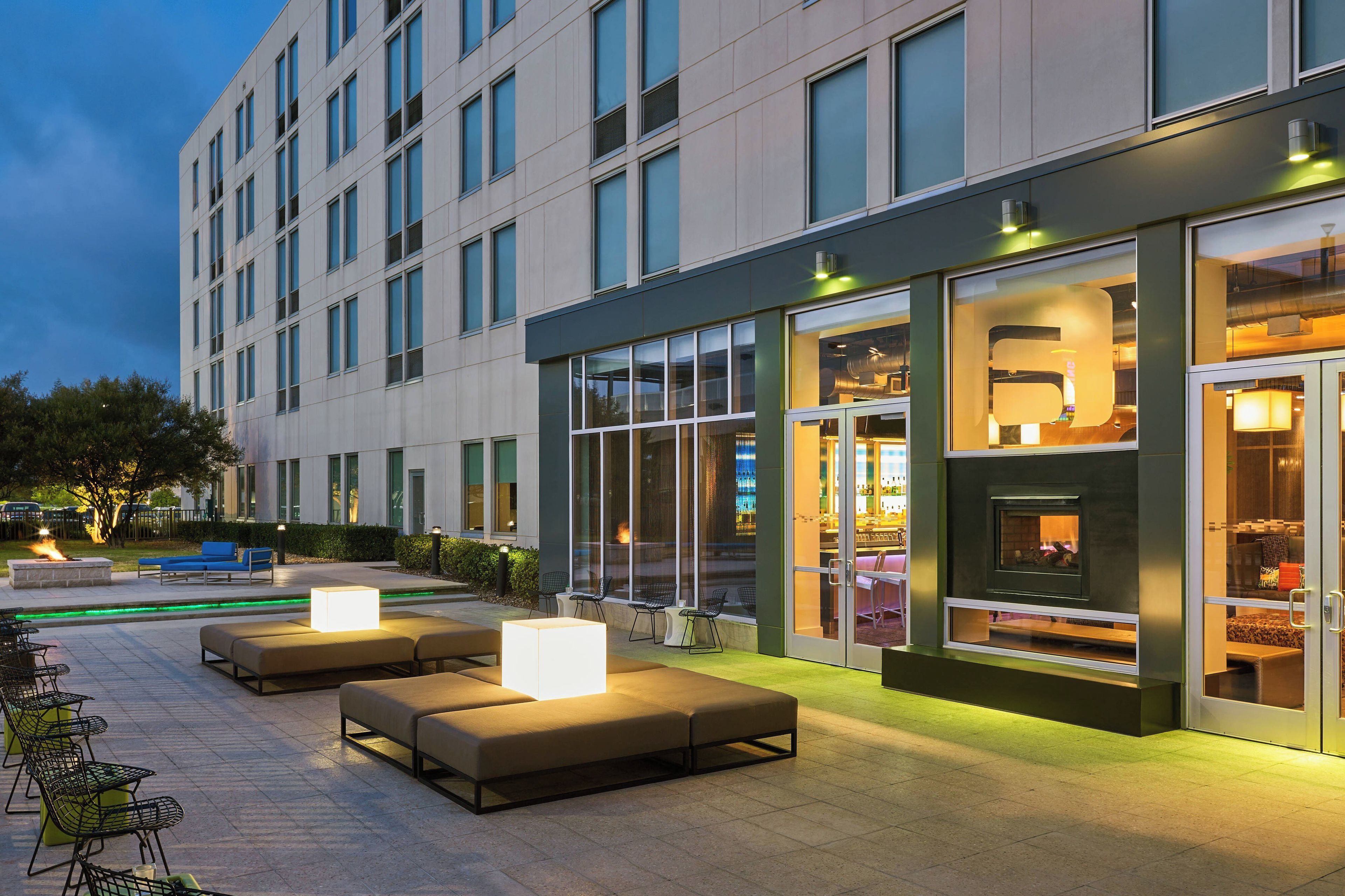 Exterior moderno de un hotel con asientos al aire libre, grandes ventanales y un bar visible.