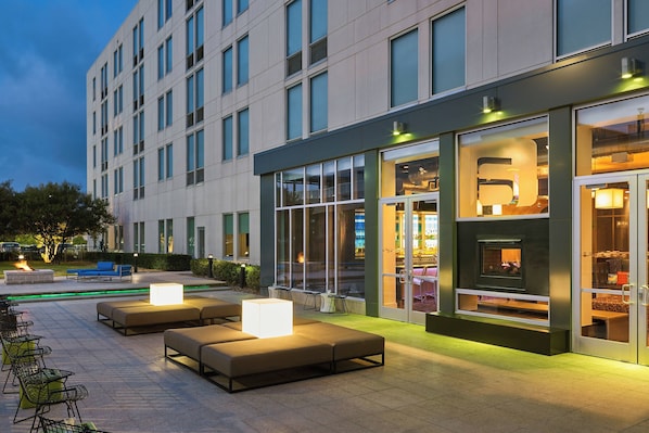 Exterior moderno de un hotel con asientos al aire libre, grandes ventanales y un bar visible.