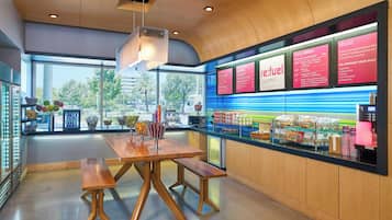 Interior moderno de café com mesa e bancos de madeira, balcão exibindo alimentos e quadros de menu.