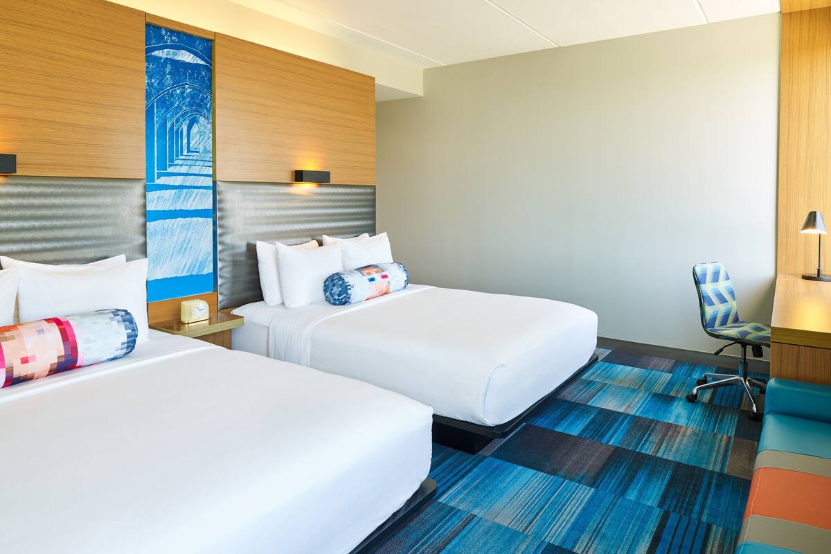 A hotel room with two beds, a desk, a chair, and a large blue artwork on the wall.
