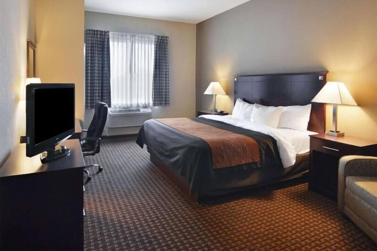 Ein Hotelzimmer mit Bett, Schreibtisch inklusive Computer, Stuhl, Fenster mit Vorhängen und zwei Nachttischlampen.