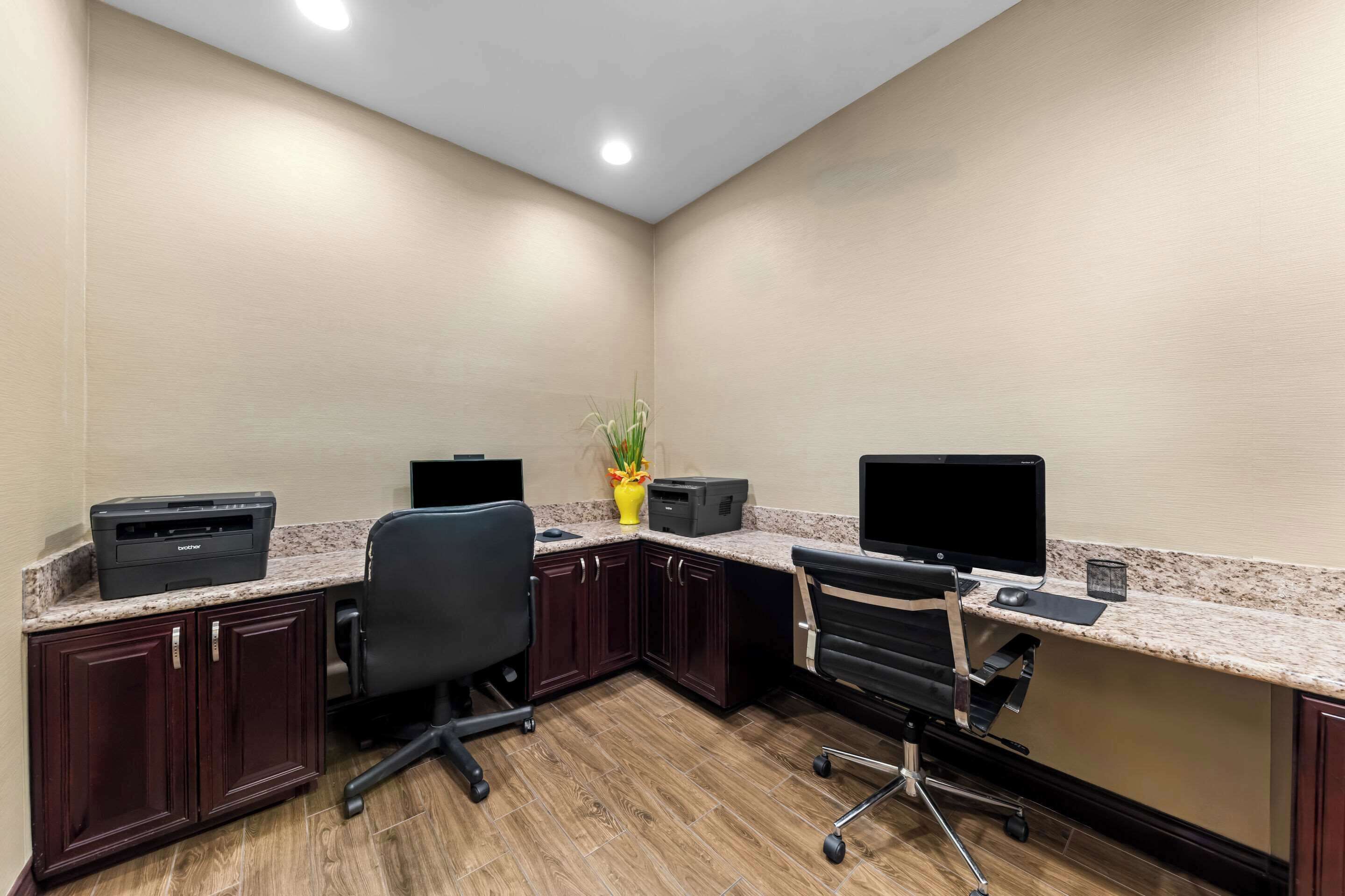 A conference room with two desks, each with a computer monitor, printer, and keyboard. There are two office chairs, a potted plant, and a small table with a lamp.