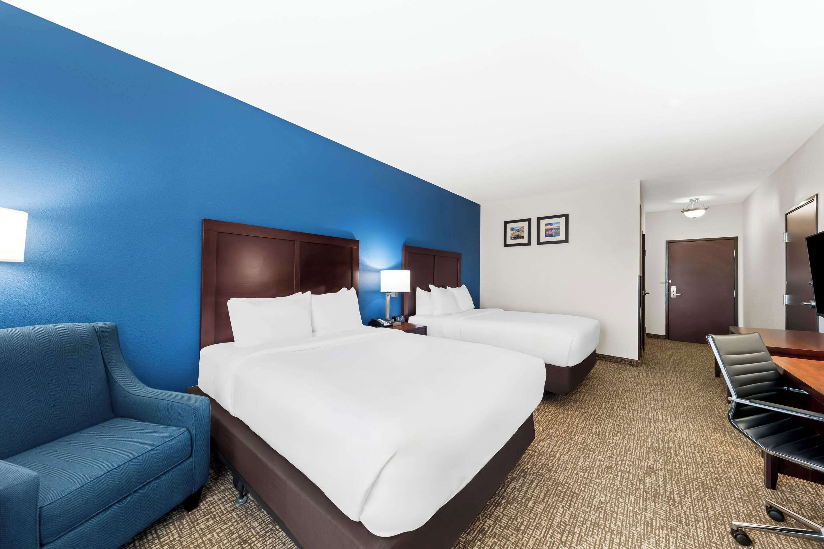 Una habitación de hotel con una cama grande, dos camas más pequeñas, un sillón azul, un escritorio con silla y una televisión.