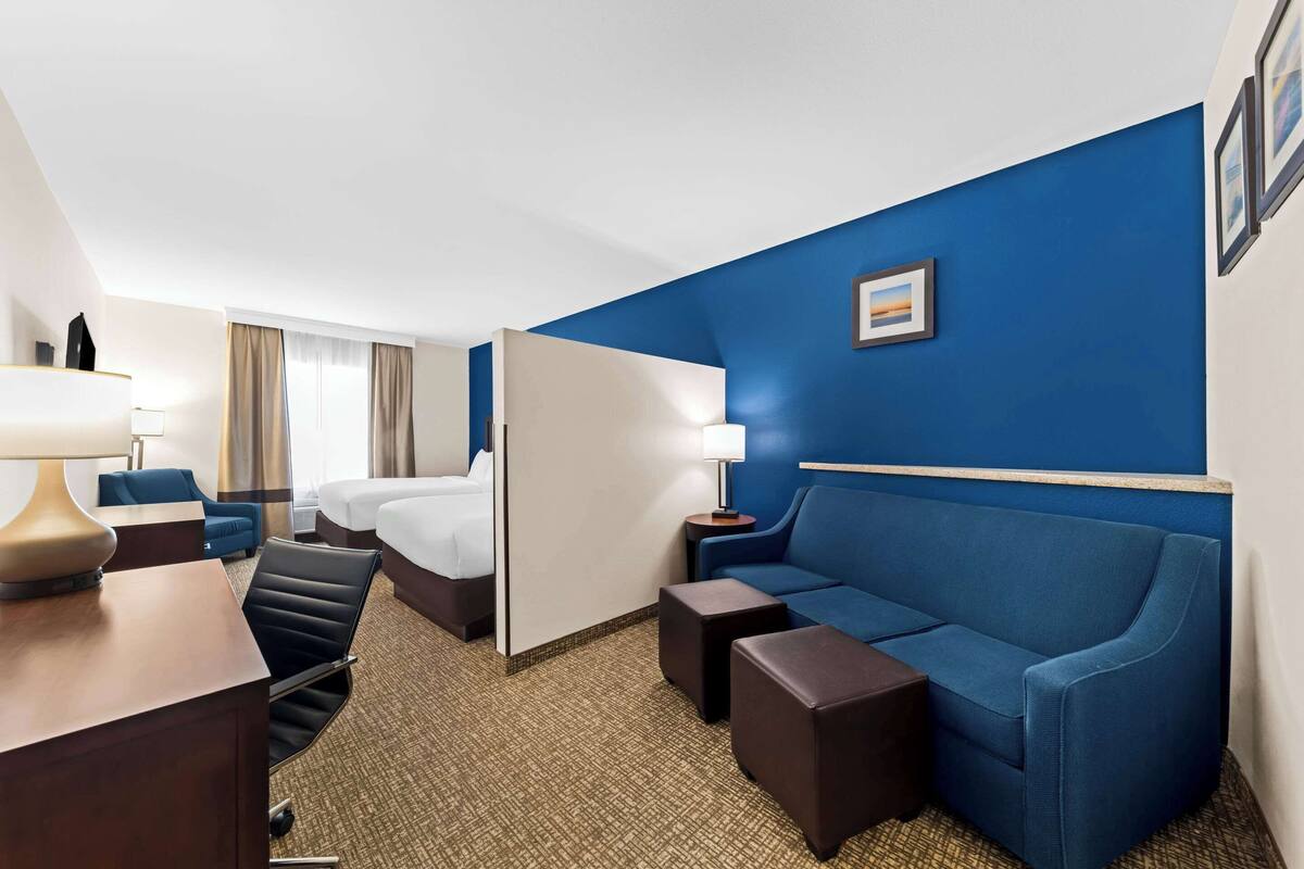 Ein Hotelzimmer mit blauer Akzentwand, einem Bett, einem blauen Sofa, einem Schreibtisch mit Lampe und einem kleinen Tisch.
