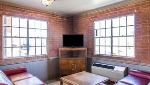 A room with brick walls, large windows, a flat-screen TV on a wooden stand, leather seating, and a table.