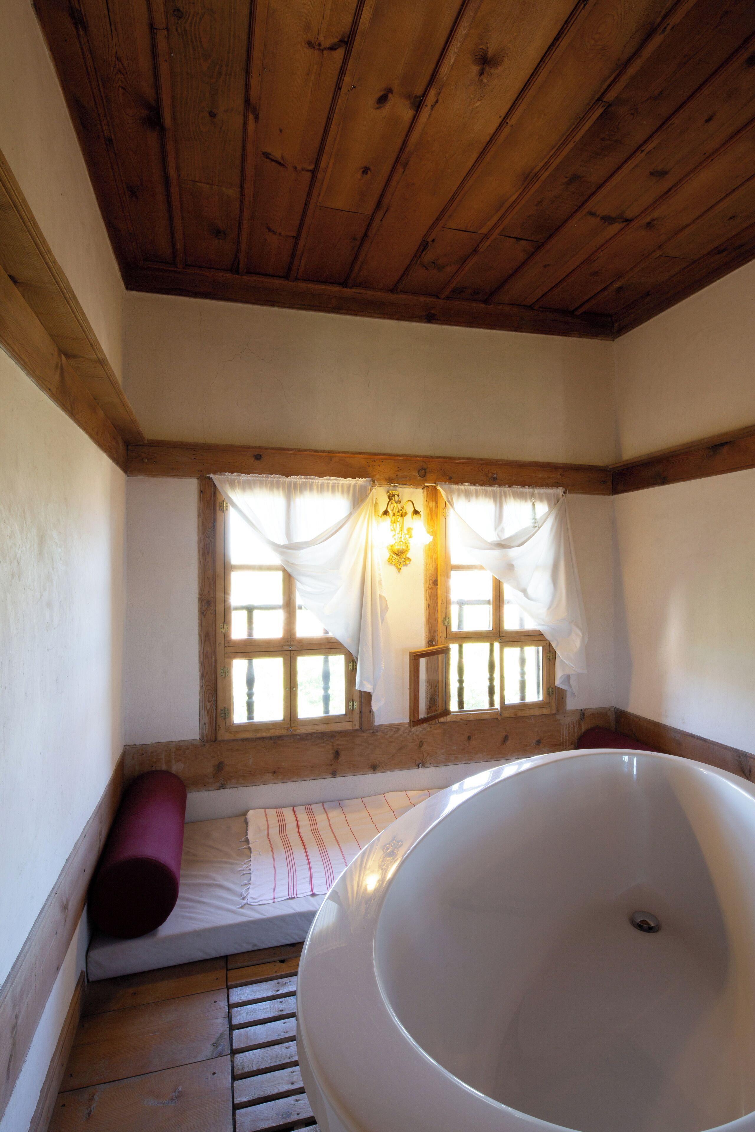 A bathroom with a wooden ceiling, a large bathtub, a window with curtains, and a wooden bench.