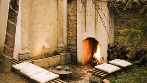 Ein Kamin aus Stein mit Feuer, eine Steinsitzbank mit Kissen und ein kleiner Tisch mit einer Schale.