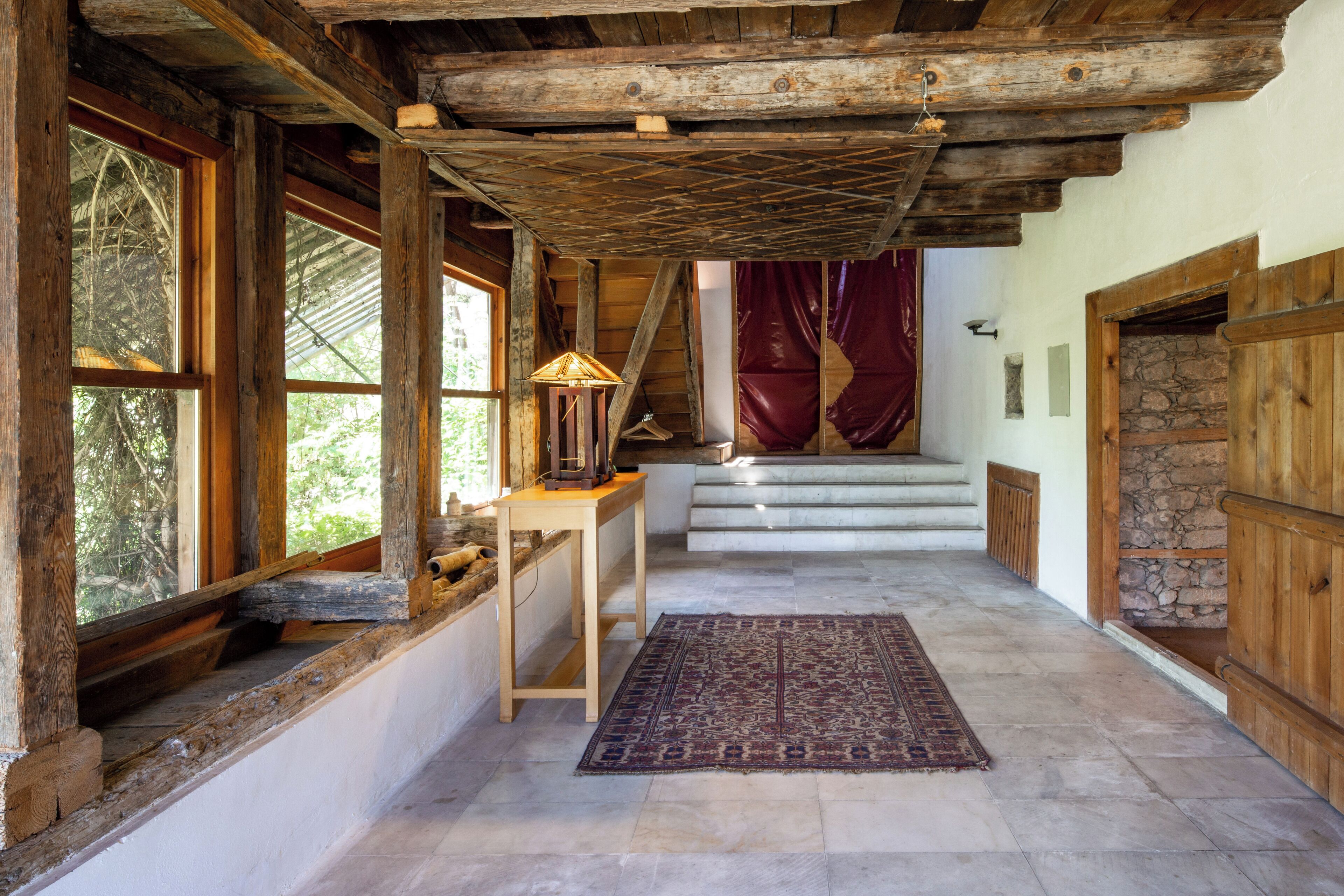 A rustic interior with wooden beams, a staircase, and a view of the outdoors.