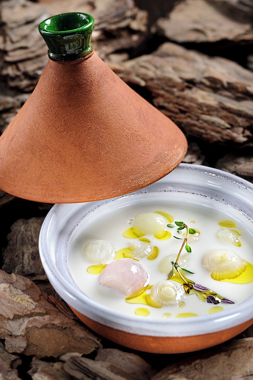Un plat traditionnel marocain, un tajine, servi dans un récipient en argile avec son couvercle, sur une assiette blanche, accompagné d’olives et d’herbes aromatiques.