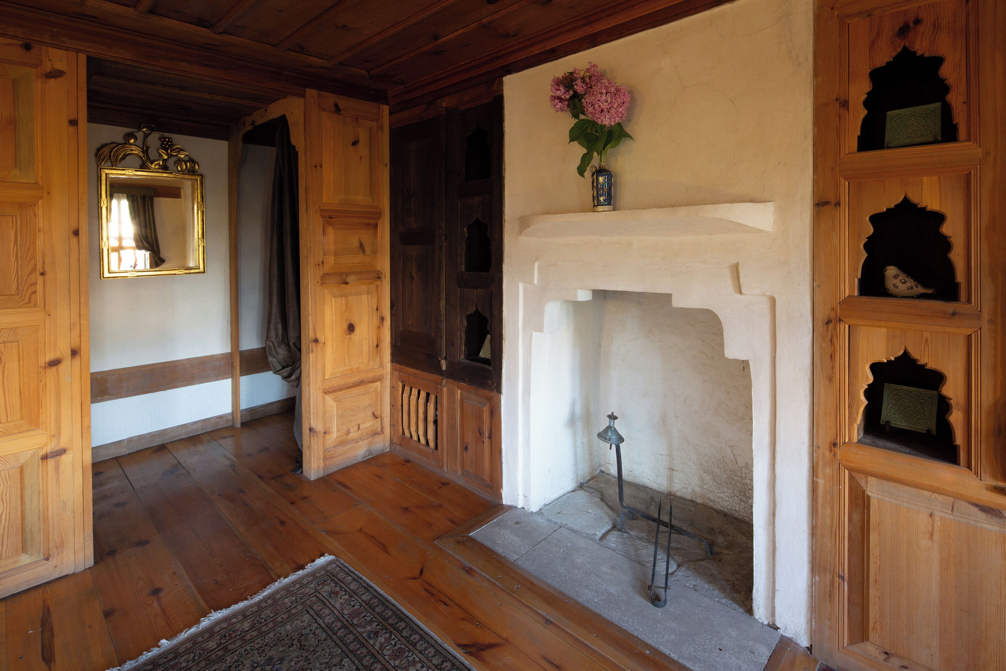 Une pièce au plancher de bois, dotée d’une cheminée, d’un miroir et d’étagères ornées d’objets décoratifs.