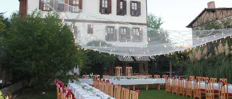 Un área de comedor al aire libre con mesas dispuestas para los comensales, rodeada de sillas de madera y un mantel blanco.