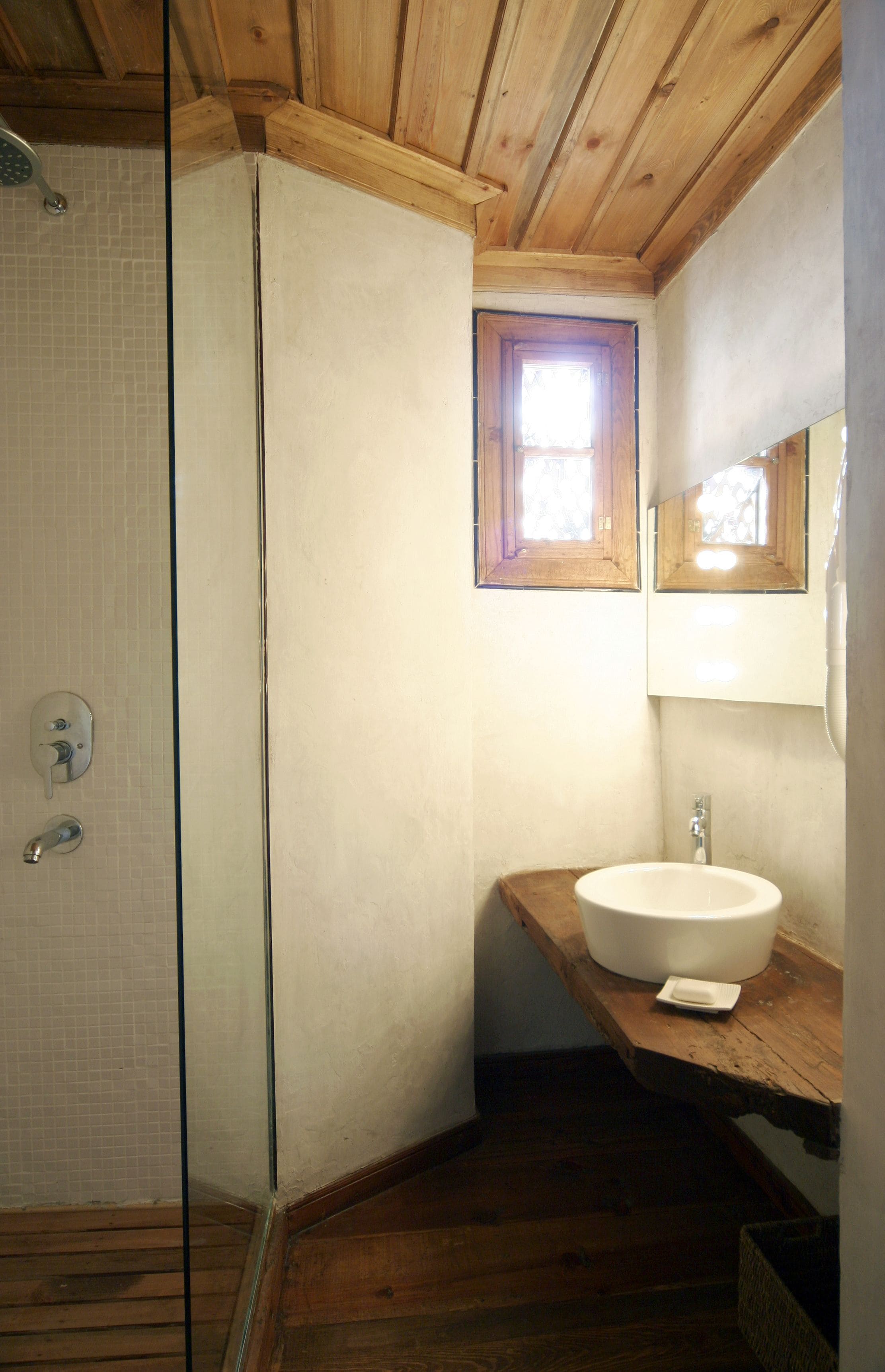 A bathroom with a wooden ceiling, a glass shower enclosure, a white sink, and a small window.