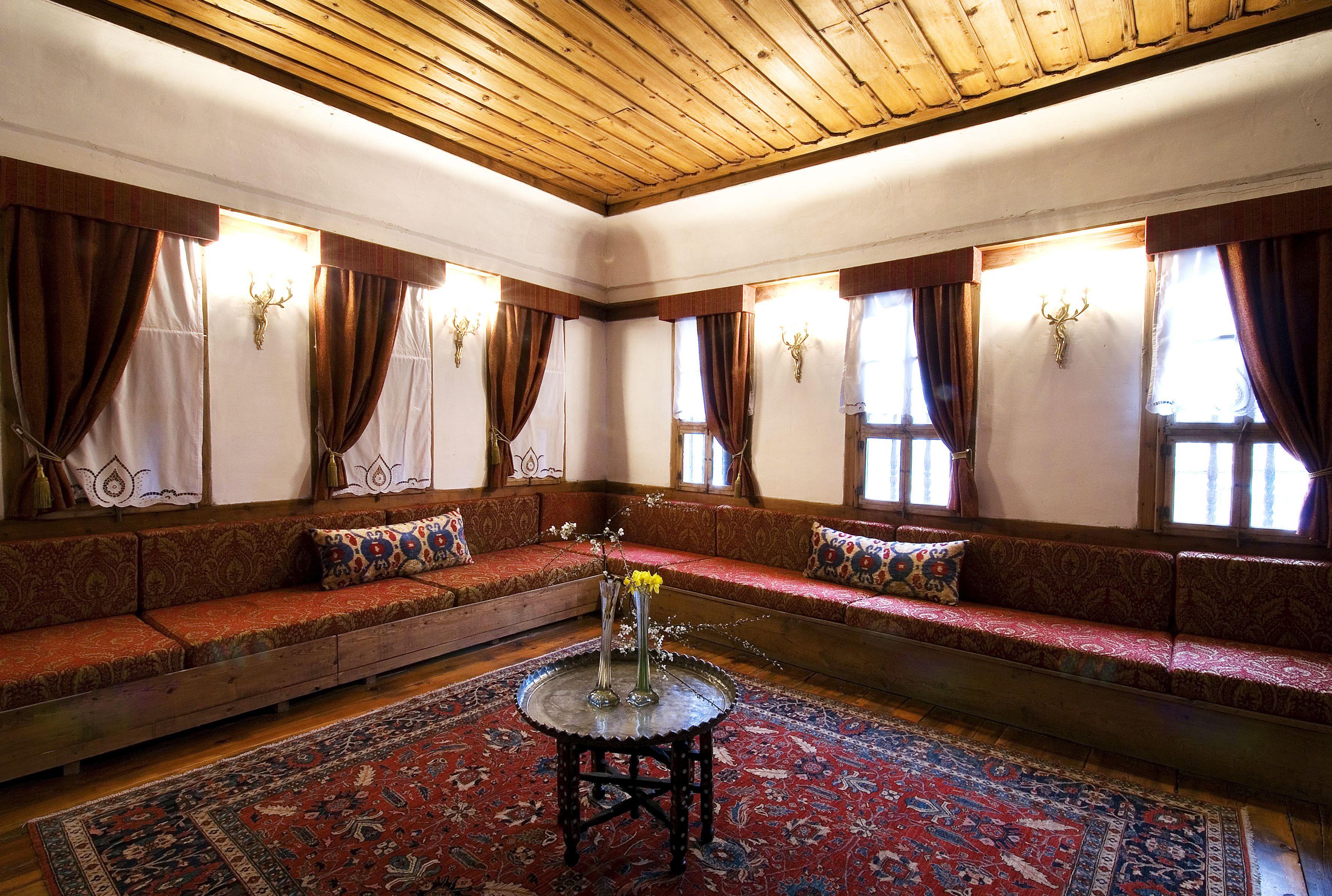 A traditional room with a wooden ceiling, patterned sofas, a central round table with a vase, and patterned rugs.