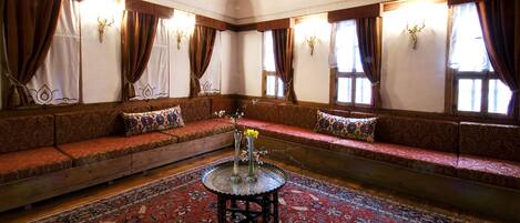 A traditional room with a wooden ceiling, patterned sofas, a central round table with a vase, and patterned rugs.